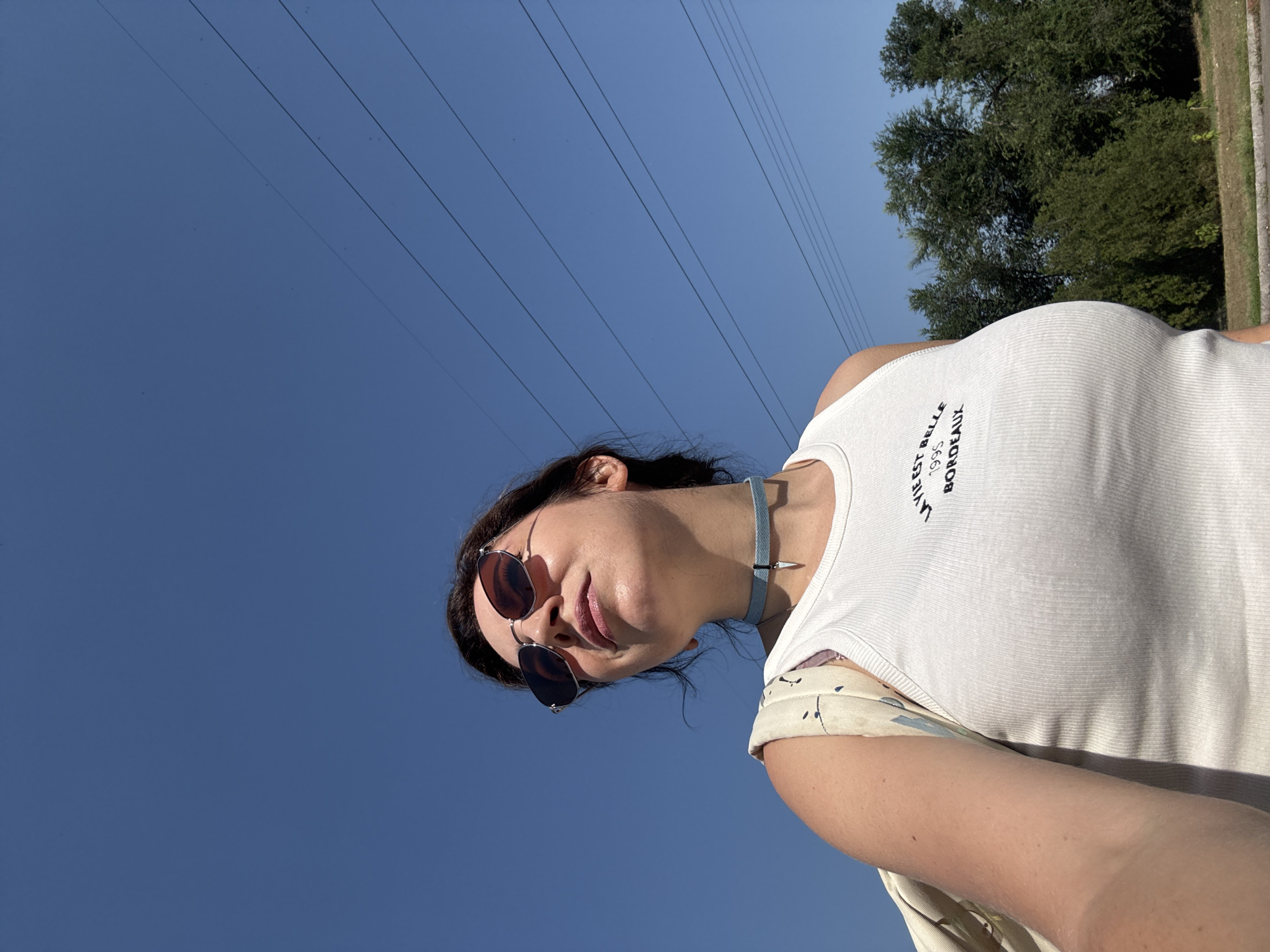 A woman is standing outside on a sunny day, looking up. She has on sunglasses, a white tank top with text that reads "LA VIE EST BELLE 1995 BORDEAUX", and a choker necklace. She is carrying a bag on her left shoulder. In the background, there are trees, power lines, and a clear blue sky. This scene is not identifiable as Kryvyi Rih, Ukraine.