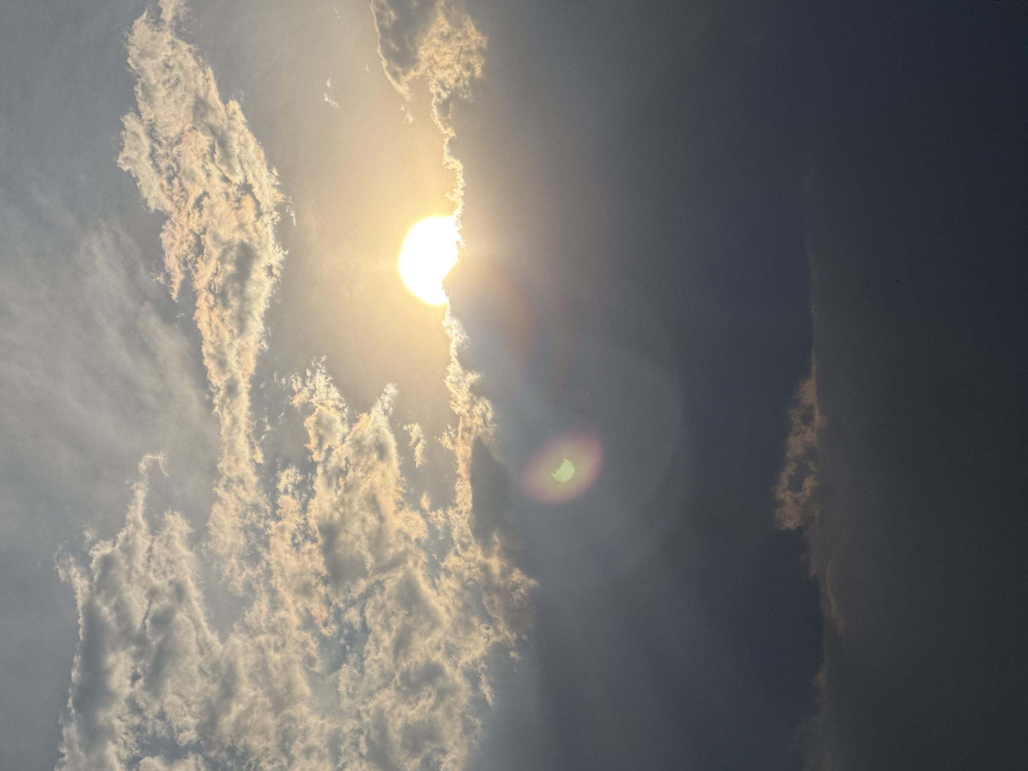 The image shows a daytime sky, seemingly in San Francisco Ocotlán, Mexico based on the prompt. The sun is partially obscured by clouds, creating a bright glare. To the left, the sky is filled with white, billowy clouds. To the right, the sky is darker, with less distinct clouds. A lens flare is visible near the center of the image.