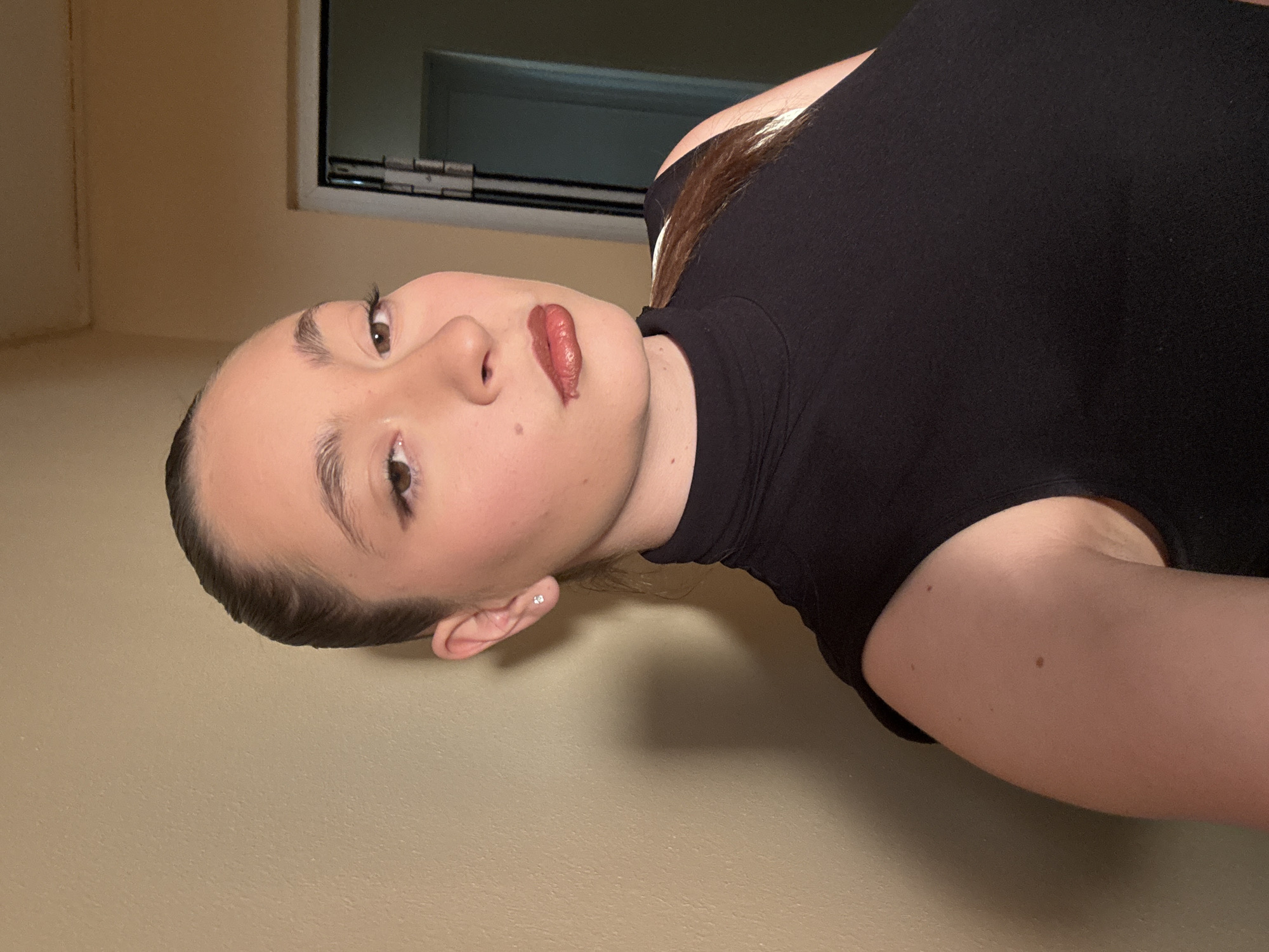 A young woman with her hair pulled back in a ponytail poses for a selfie indoors, looking towards the camera with a neutral expression. She is wearing a black sleeveless turtleneck top. Her makeup includes winged eyeliner and brown lipstick with a glossy finish. She has a small stud earring in her visible ear. The background is a plain, light beige wall, with a doorway and part of a doorframe visible to her right. The lighting appears to be artificial, casting a shadow to her left. The image does not provide any information about the city of Sofia, Bulgaria.