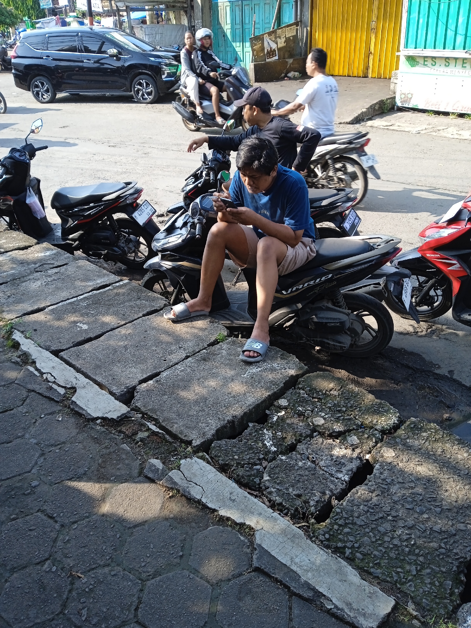 The image depicts a street scene in Kaliwungu, Indonesia. A young man in a blue shirt and khaki shorts sits on a black and silver "Vario" motorcycle, looking at a smartphone. Several other motorcycles are parked nearby, including black and red Honda motorcycles. Two men are on another motorcycle behind him and a third is walking in the same direction. A black SUV is parked on the other side of the road. A cracked concrete sidewalk borders the parking area. Behind the parking area are shops and buildings.