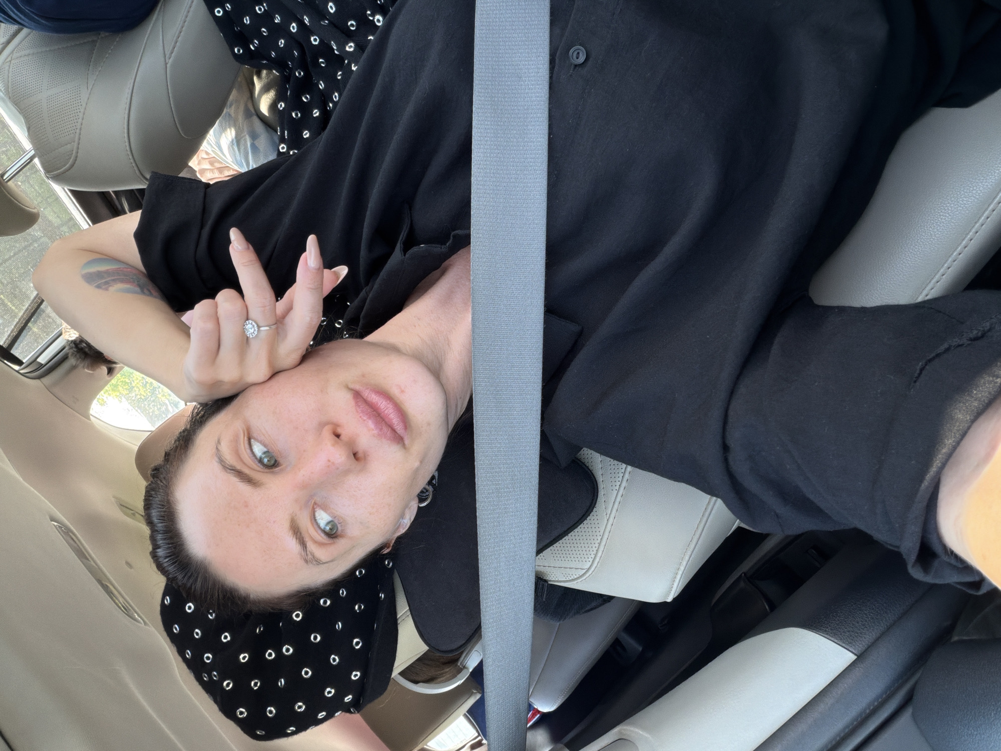 A woman is lying down in the backseat of a car, with her head resting on a pillow. She is wearing a black shirt and has her hand raised to her face. The car appears to be moving, as suggested by the seatbelt across her chest. The interior of the car is visible, with beige leather seats and the car's ceiling. The woman has fair skin, dark hair, and is looking away from the camera.