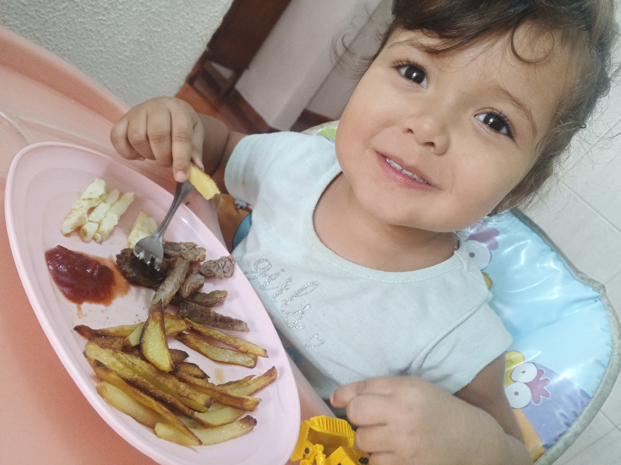 An indoor, eye-level close-up photograph captures a young child, likely a toddler, seated in a high chair during a meal. The child, with light brown skin, dark eyes, and curly brown hair, looks directly at the camera with a cheerful, open-mouthed smile, revealing small teeth. They are wearing a light blue short-sleeved t-shirt with the word "girly" subtly visible in sparkly letters on the front.
In their left hand, the child holds a metal fork, which has speared a piece of food, possibly a potato wedge. A pink rectangular plate is on the high chair's tray, laden with a meal consisting of golden-brown French fries or potato wedges, several pieces of cooked meat (appearing to be beef or similar), a dollop of red ketchup, and some lighter-colored, cut food items, possibly cheese or another type of potato. The child's right hand rests near a yellow toy truck, which is also on the tray.
The high chair features a blue and purple patterned cushion with cartoon bird or chicken illustrations. The background provides glimpses of an indoor setting with light-colored walls and furniture, typical of a home in Caracas, Venezuela, where this scene takes place. The image conveys a happy and engaged mealtime.