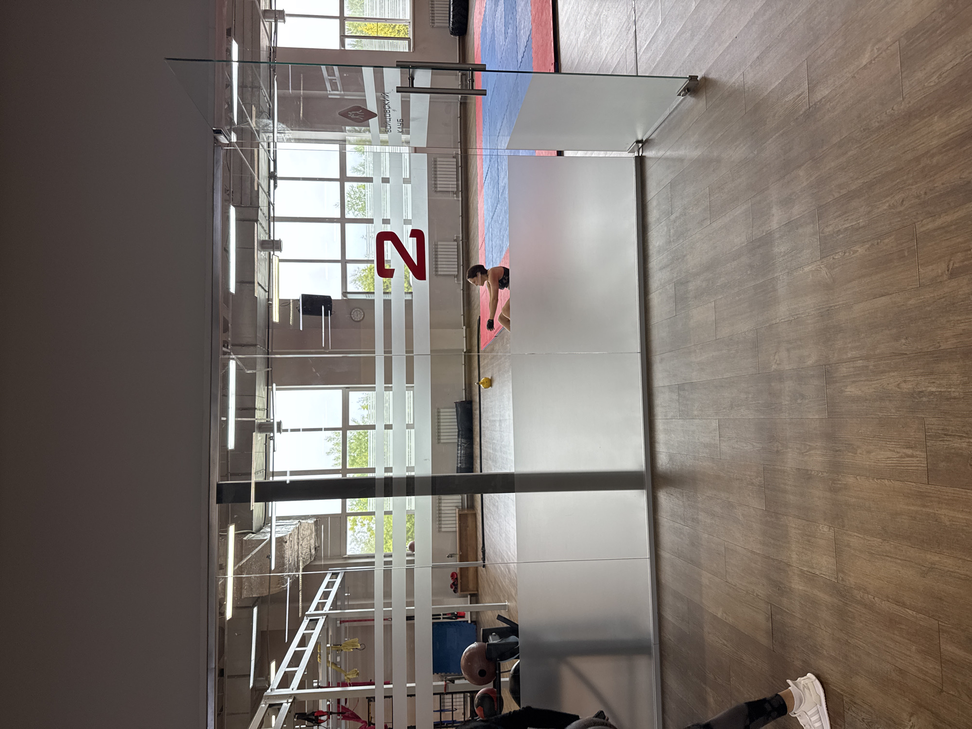 An indoor shot captures a gym with a glass partition and wooden flooring. The partition has frosted horizontal stripes and a large red number "2" on it. Inside the gym, visible through the glass, a person is performing sit-ups on a mat. The gym area is equipped with various training apparatus, including a large tire, mats on the floor in red and blue, and a wooden bench. Large windows offer a view of trees and the sky outside. The overall impression is a clean, modern fitness facility. The image does not contain any specific visual cues to pinpoint the exact location in Chaikovsky, Russia, beyond it being an indoor sports facility.