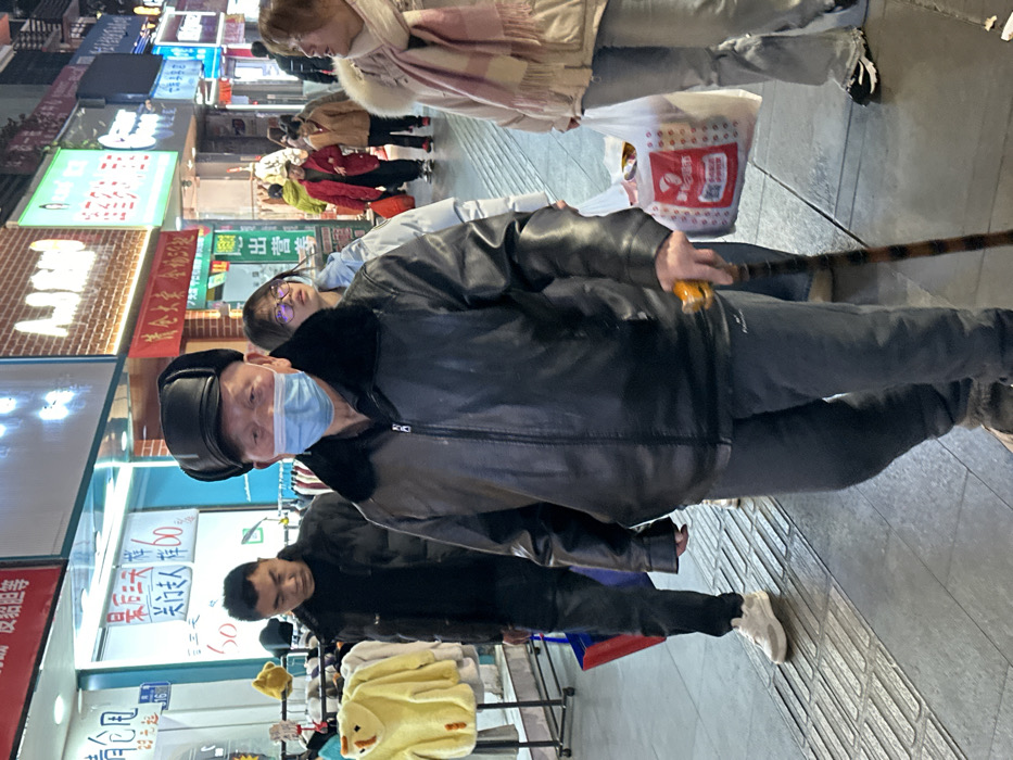 A nocturnal scene depicts a commercial street in Jiangjin, China, illuminated by multiple storefront signs. In the foreground, an elderly Asian male walks towards the left, wearing a black leather jacket with a fur collar, a black winter cap with ear flaps, grey trousers, and a light blue surgical mask. He holds a brown wooden walking stick with an orange handle in his right hand. Behind him, a younger Asian male in a black jacket and dark pants carries a blue shopping bag. To his right and slightly behind, a young Asian female with glasses wears a light-colored puffer jacket. Further right, another Asian female, dressed in a light-colored coat and a pink striped scarf, carries a white plastic shopping bag with a red logo. The street features a tiled sidewalk with tactile paving strips. Storefronts display brightly lit signs with Chinese characters and some English text, showcasing clothing merchandise such as a yellow fuzzy garment and various hats.