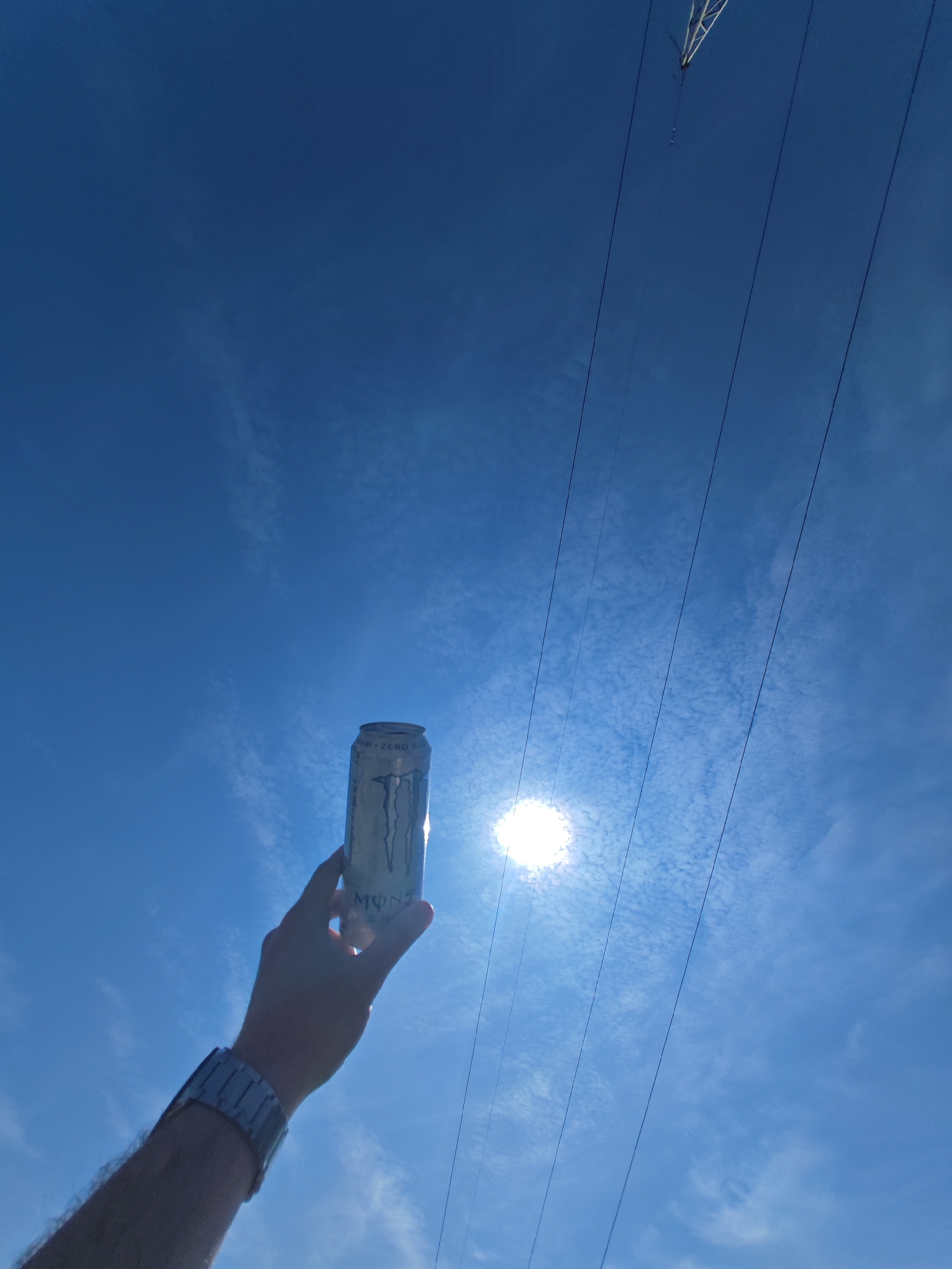 The image depicts an outdoor scene in the city of Shumen, Bulgaria, observed from a low-angle perspective looking upwards towards a bright blue sky. Scattered wispy white clouds are visible across the sky. The sun is a prominent, overexposed light source positioned in the lower-right quadrant. A pale-skinned human forearm and hand, with visible arm hair, extends from the lower-left into the frame. The wrist wears a silver-colored metal link watch. The hand is gripping a silver-colored cylindrical beverage can, identified as a "MONSTER ZERO SUGAR" energy drink, held upright, partially obscuring the sun. Several dark, taut lines, consistent with overhead power or communication cables, traverse the sky diagonally from the mid-left to the upper-right. A white, angular structural element, indicative of an electrical pylon or transmission tower, is partially visible in the upper-left corner, from which some of these lines appear to extend.