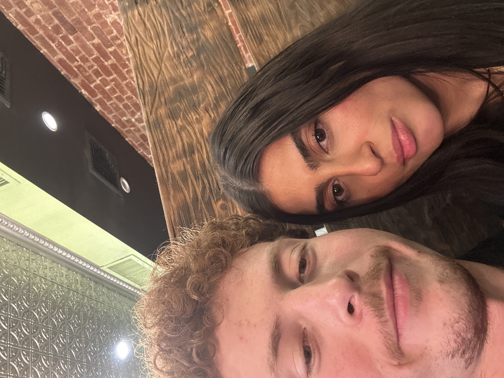 An indoor, close-up photograph features two individuals, a male and a female, positioned in a selfie-style composition. The male, on the left, exhibits light skin, reddish-brown curly hair, and a light mustache with stubble on his chin. He is looking slightly downwards with a subtle upward curve to his mouth. The female, on the right, has dark, straight hair, olive skin, and neutral-toned lips with pink lipstick; she looks directly forward with a neutral expression.
The background reveals interior architectural elements consistent with a renovated urban space, such as a restaurant or bar, within the city of New York, United States of America. Visible in the upper left is an ornate, silver-colored pressed tin ceiling with a repetitive circular motif. To the upper right, a section of an exposed red brick wall is present. A significant portion of the background on the right and center consists of dark, rustic wooden paneling with horizontal grain. Overhead, a dark ceiling or beam includes two recessed circular light fixtures and two rectangular HVAC vents. A light green horizontal architectural element or painted beam runs above the male's head.