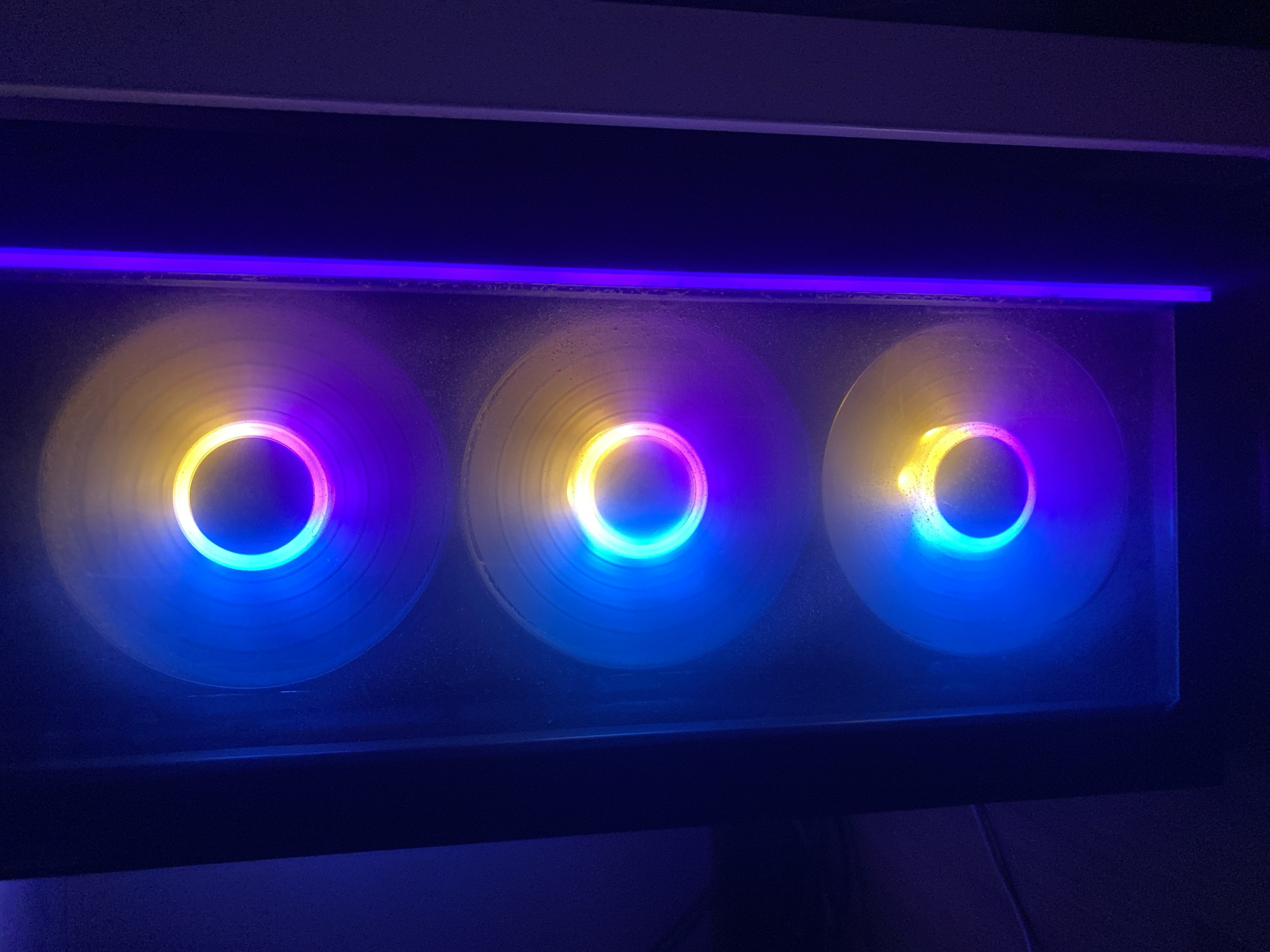 The image shows a close-up view of three computer case fans illuminated with RGB lighting. The fans are mounted horizontally, likely in a computer case. The RGB lighting displays a color gradient transitioning from yellow near the center to blue on the outer edges of the fan blades, with a ring of purple separating the yellow and blue hues. The fans are situated behind a transparent panel, possibly made of glass or acrylic, which appears slightly dusty or smudged. A purple LED strip is visible above the fans. The overall environment is dark, with surrounding surfaces appearing black or dark blue. The location (Budva, Montenegro) cannot be determined from the provided image.