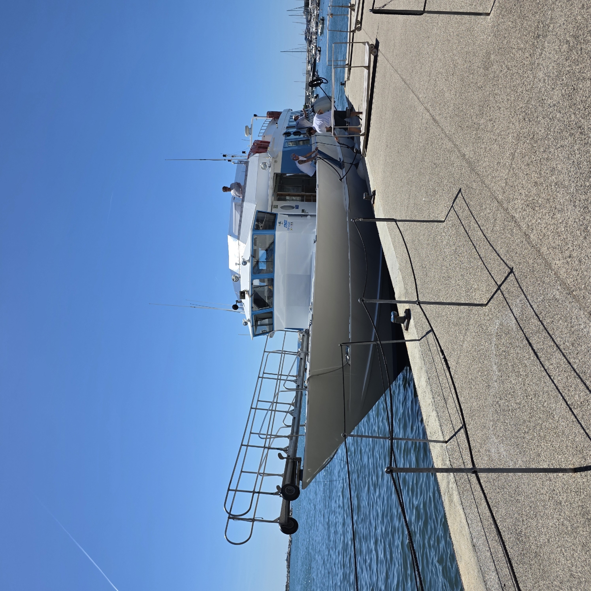 The image depicts a white passenger boat docked at a waterfront promenade in Rapallo, Italy. The boat is long and sleek, with a prominent white superstructure and blue-tinted windows. Several people are visible on and around the boat. Two individuals are on the deck of the boat, one near the rear and another standing on the roof of the cabin. Three other individuals are on the dock, attending to the mooring lines of the boat, with one person appearing to hand a rope to another. The promenade is paved with a coarse, light-colored aggregate material, and a metal railing with black ropes separates the promenade from the water. The water is calm and blue, reflecting the clear, bright blue sky. In the background, other boats, including sailboats and smaller vessels, are moored in a harbor. The scene suggests a sunny day with maritime activity.