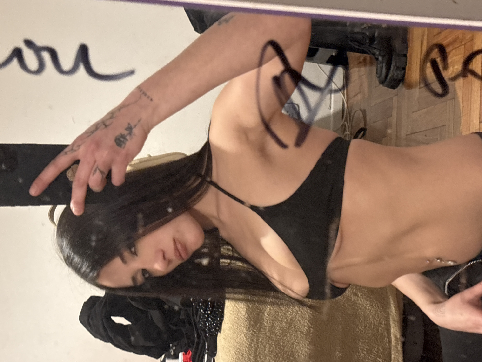A woman in a black bra is posing in front of a mirror, taking a selfie with her phone. She has dark hair and tattoos on her arm and hand. The background shows a room with a wooden floor and various objects, including what appears to be a jacket and possibly shoes. There is writing on the mirror. The location cannot be determined without further information.