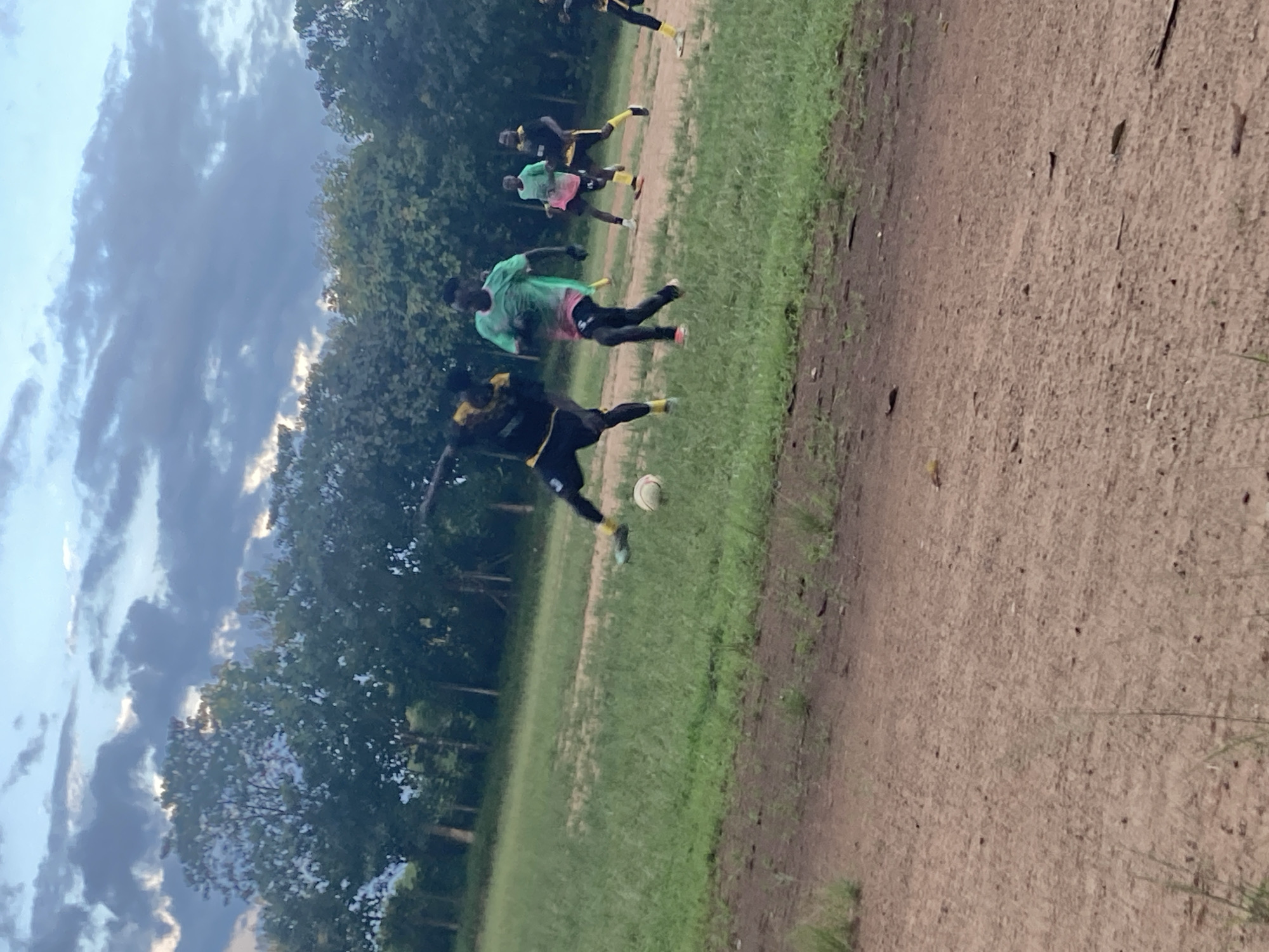 The image depicts an outdoor scene with a group of young men playing soccer on a dirt and grass field. The sky above is partially cloudy with dramatic dark clouds and hints of blue sky. In the background, a line of trees forms a dark silhouette against the sky.
In the foreground, a dirt path is visible, with sparse grass growing on it. The field itself is a mix of green grass and bare dirt patches.
Several young men, appearing to be in their late teens or early twenties, are actively engaged in the soccer game. One player in the foreground is wearing a black and yellow jersey and black shorts, mid-stride, with his right foot extended towards a white soccer ball. Another player, wearing a bright green jersey with pink accents and black pants, is running towards the ball. Further back, two other players are visible, one in a yellow and black jersey and the other in a similar green jersey to the second player. Another player is visible further in the background on the right side of the frame, also in a yellow and black jersey.
The overall impression is a casual soccer game taking place in a rural or semi-rural setting, possibly on the outskirts of Miango, Nigeria, given the vegetation and open field. The lighting suggests it may be late afternoon or early evening, with the sun low on the horizon.