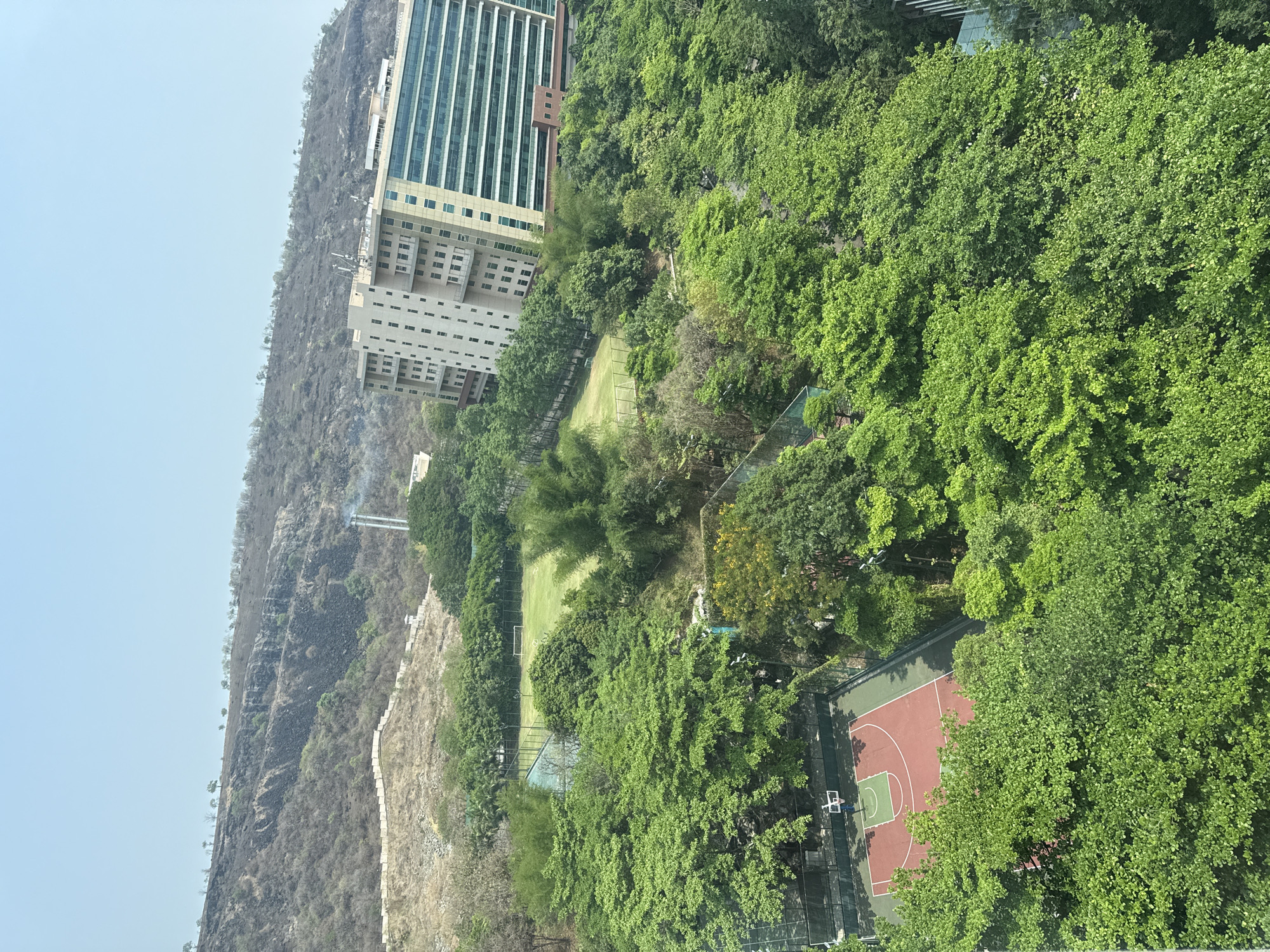 The image shows a landscape in or near Man, India. A multi-story building stands near the base of a mountain. Lush greenery surrounds the building. There's a sports field and a basketball court among the trees. The sky is hazy.