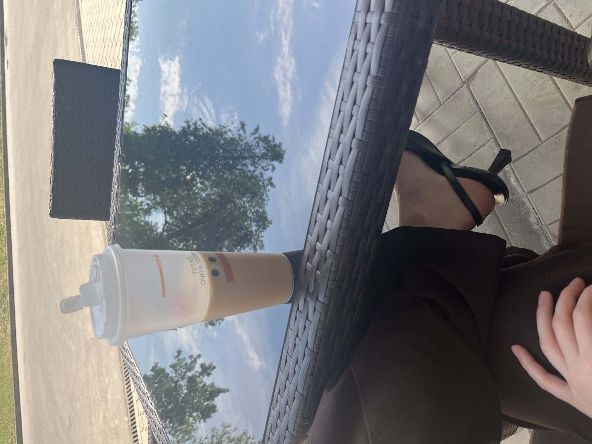 A high-angle shot shows a person sitting at an outdoor, dark-brown wicker table. A partially-filled plastic cup sits on the table. The cup has Russian text reading "ТЕПЛО ОЧЕНЬ ТЕПЛО" (warm very warm). The table's glass surface reflects trees and a bright sky with scattered clouds. The person is wearing dark brown pants and a black heeled sandal. Their right hand rests on their lap. The setting appears to be outdoors, possibly in Moscow, Russia.