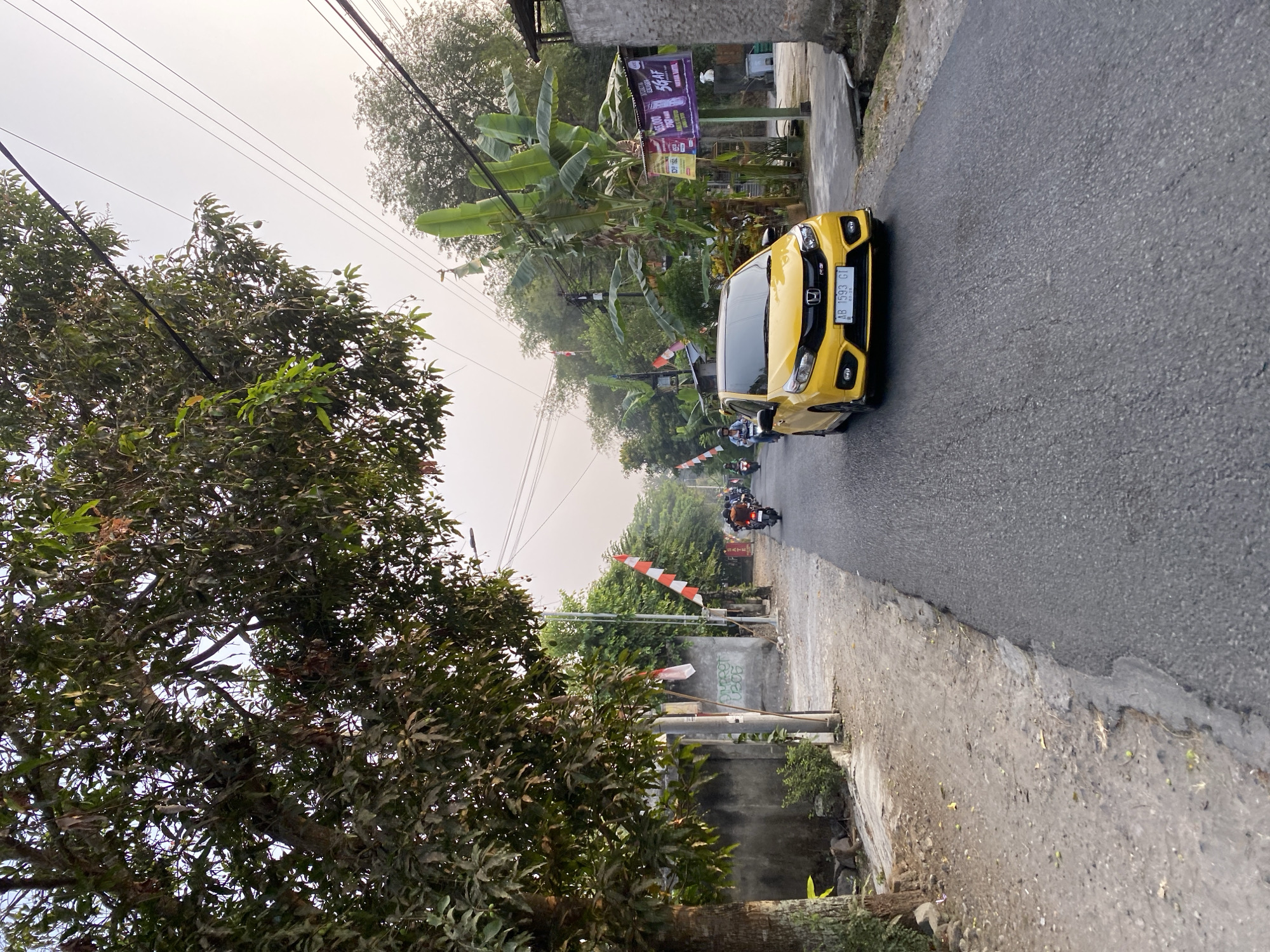 A sunny day view captures a residential street in Kasihan, Indonesia. A bright yellow car with license plate AB 1593 GI drives down the asphalt road. To the left of the road, a mature tree with dark green leaves shades a dusty path and a low concrete wall. Red and white Indonesian flags are strung along the wall, indicating a national celebration. In the distance, several motorcycles and their riders are seen on the road. Banana trees and other lush vegetation line the right side of the street, with banners advertising mobile phone services hanging from a utility pole. The sky is a pale, hazy blue, suggesting it might be late afternoon or early morning.
