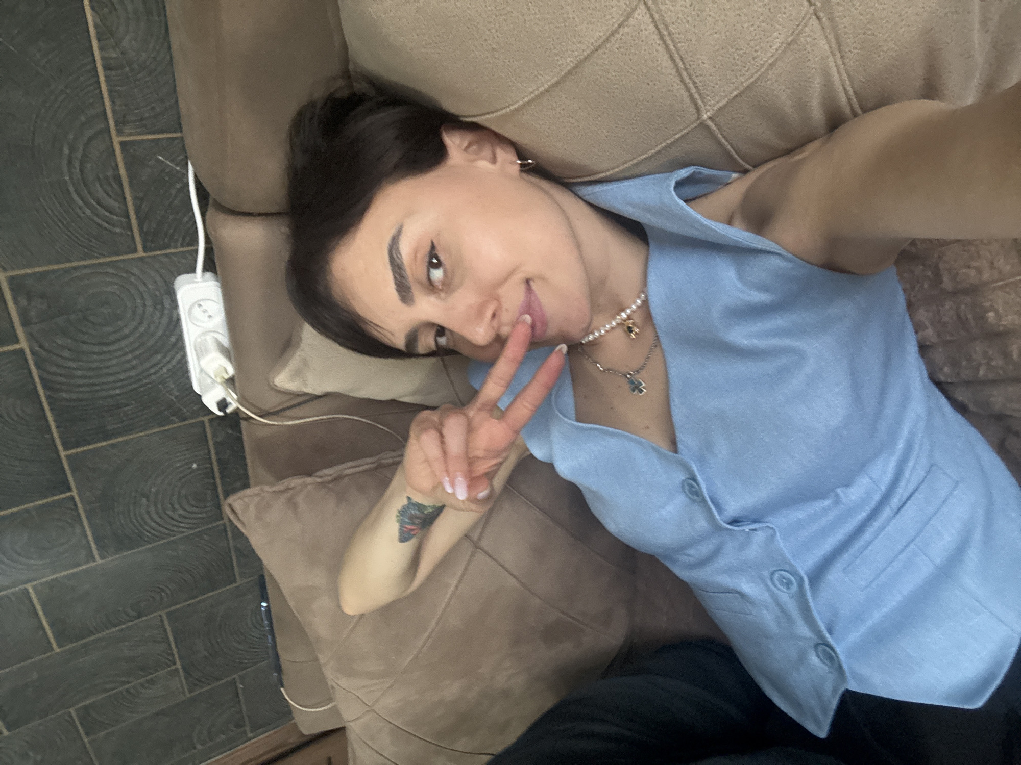 A woman is lying on a brown sofa, making a peace sign. She is wearing a light blue button-down vest, a pearl necklace with a pendant, and has a tattoo on her left forearm. An electrical outlet and cord are visible on the floor next to the sofa. The floor is tiled with dark grey rectangular tiles that have a wood-grain pattern. The location is not identifiable as Tbilisi, Georgia from the visual elements in the image.