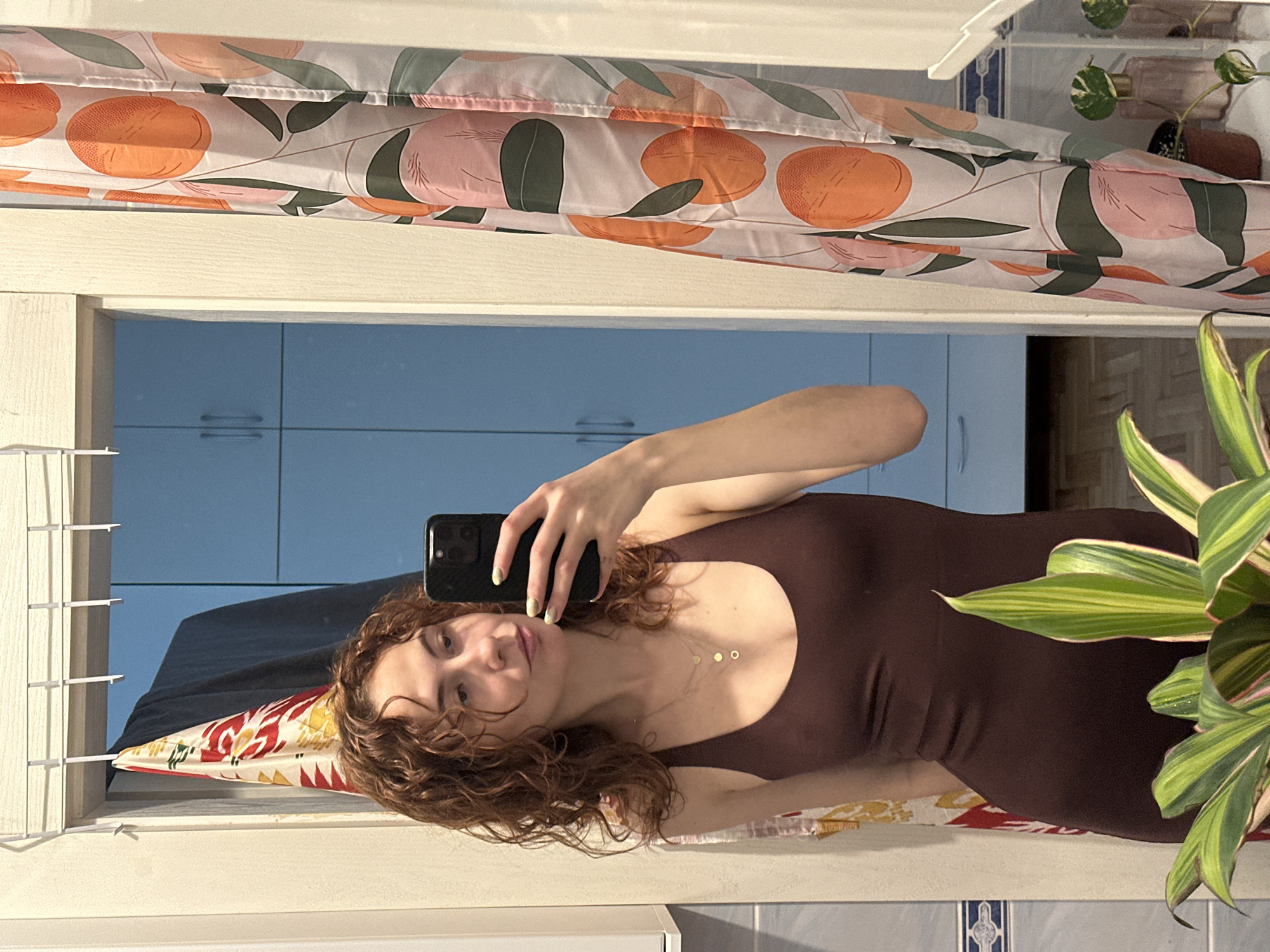 A woman is taking a selfie in a bathroom mirror. The woman has curly brown hair and is wearing a brown dress and a gold necklace. She is holding a black smartphone in her right hand. The mirror reflects a bathroom with blue cabinets. A shower curtain with an orange fruit pattern hangs above the mirror. A plant is visible in the bottom right corner. The exact location in Novi Sad, Serbia cannot be determined.