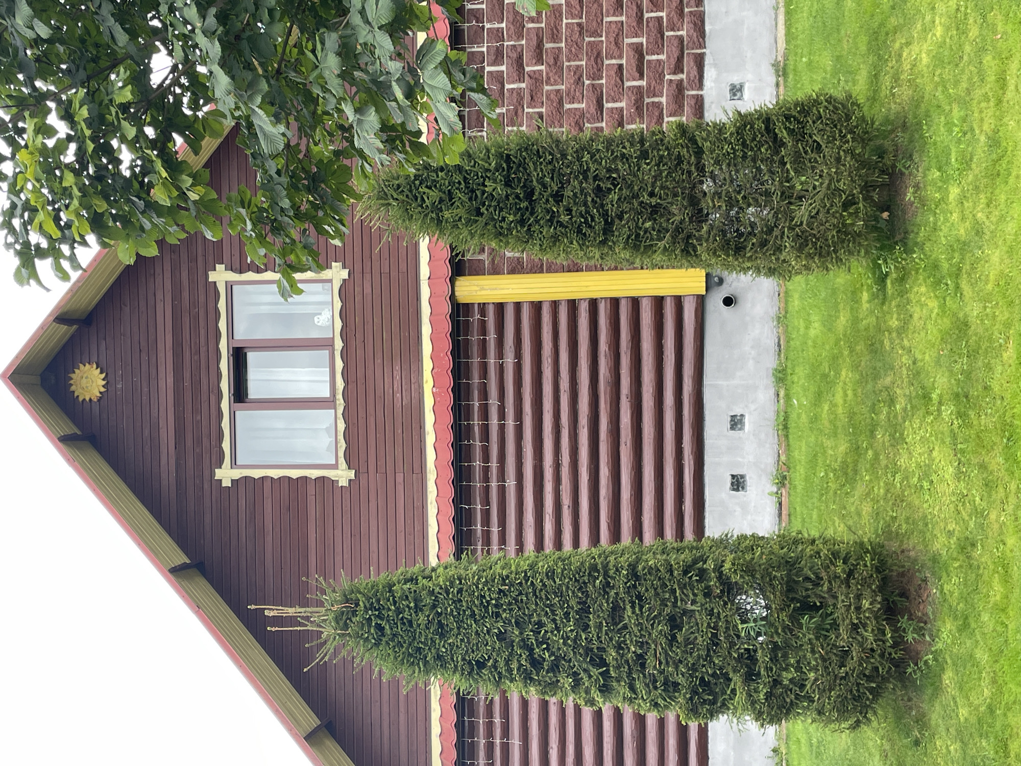 The image depicts the exterior of a rustic, two-story wooden house located in the city of Строймаш Снт, Russia. The house features dark brown horizontal log siding on the lower portion and dark brown plank siding on the upper gable. A decorative yellow trim outlines the window in the gable, which has a sun-shaped ornament above it. The roofline is accented with a red and yellow trim. Below the upper window, a section of the house is covered in red brick siding. Two tall, conical evergreen trees stand in the foreground on a well-maintained green lawn. The sky is overcast.