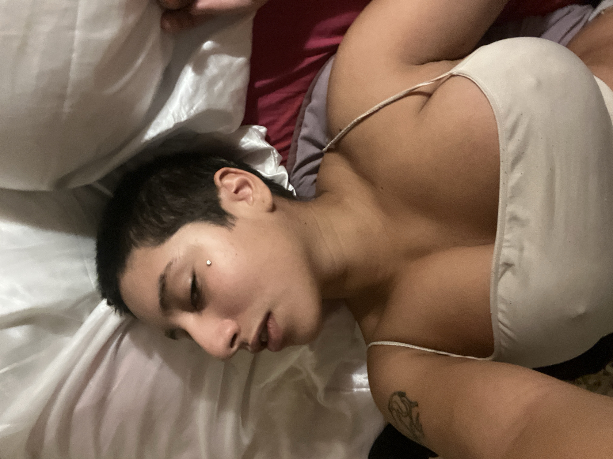 A person with short dark hair is lying on their back on a white satin pillow and white satin sheets. They are wearing a light-colored top with thin straps. A tattoo is visible on their left arm. A small stud earring is in their left eyebrow. A red-colored pillow is visible behind the person. The person appears to be resting. The location is not discernible from the provided image.