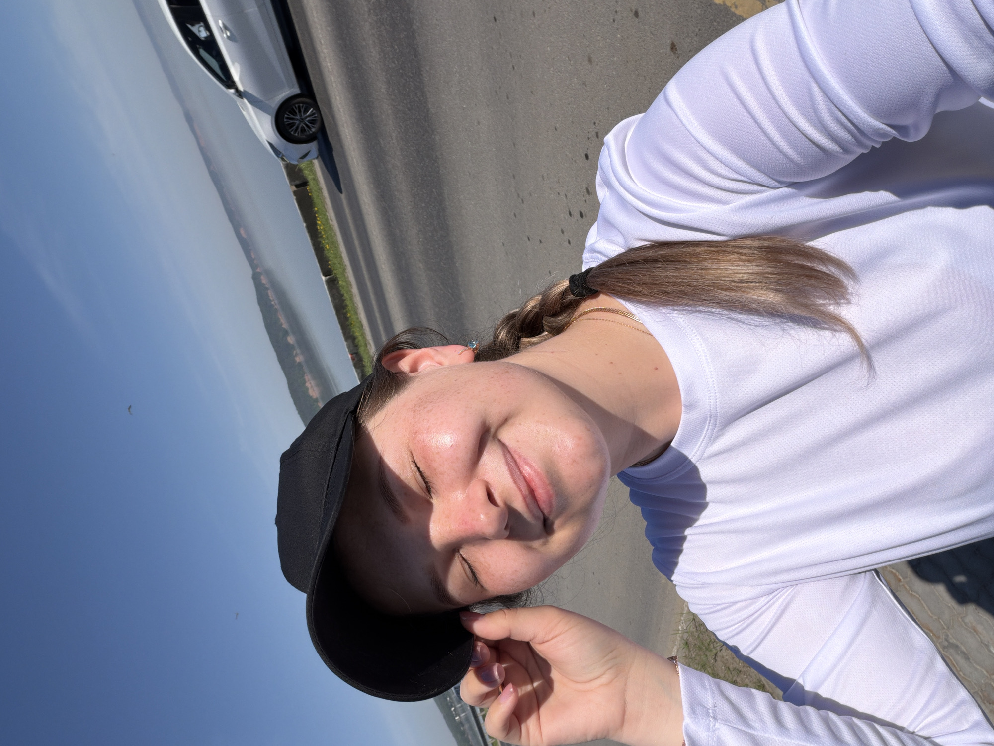 A woman with a braid and a black baseball cap is smiling with her eyes closed on what appears to be a road. A white car is parked to the left of her. In the background, there is a landscape of trees and water under a blue sky. This image was taken in Chaikovsky, Russia.