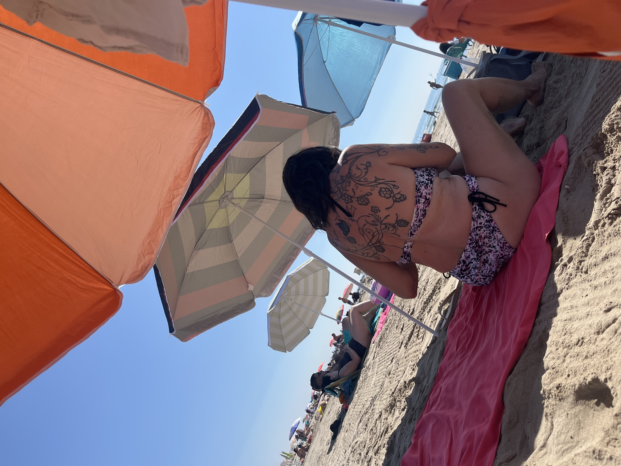The image captures a sunny day on the beach in Palavas-les-Flots, France. The foreground features a woman sitting on a pink towel, facing away from the camera, with a floral tattoo on her back. She is wearing a patterned bikini and is partially shaded by an orange umbrella. To her right, a blue umbrella provides shade, and an orange garment is draped over its pole. In the background, other beachgoers are seen relaxing under umbrellas, and the sea is visible in the distance. The sand is the primary element of the environment, with a clear blue sky overhead.