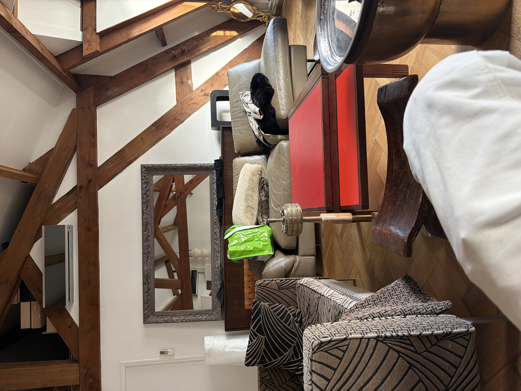 The photograph depicts an interior space, possibly in Paris, France, given the architectural details often associated with Parisian interiors. The room features exposed wooden beams characteristic of attic or loft conversions. Dominating the scene is a collection of stacked furniture and objects, including upholstered cubes with a brown and black geometric pattern, a stack of tan leather seating cushions, a red panel, a large silver bowl, and other miscellaneous items. A framed mirror reflects a view of the structural beams. A bright green shopping bag is visible among the stacked objects. The presence of packing materials suggests that the space may be in the process of being organized or rearranged. The overall impression is of a cluttered or transitional space.
