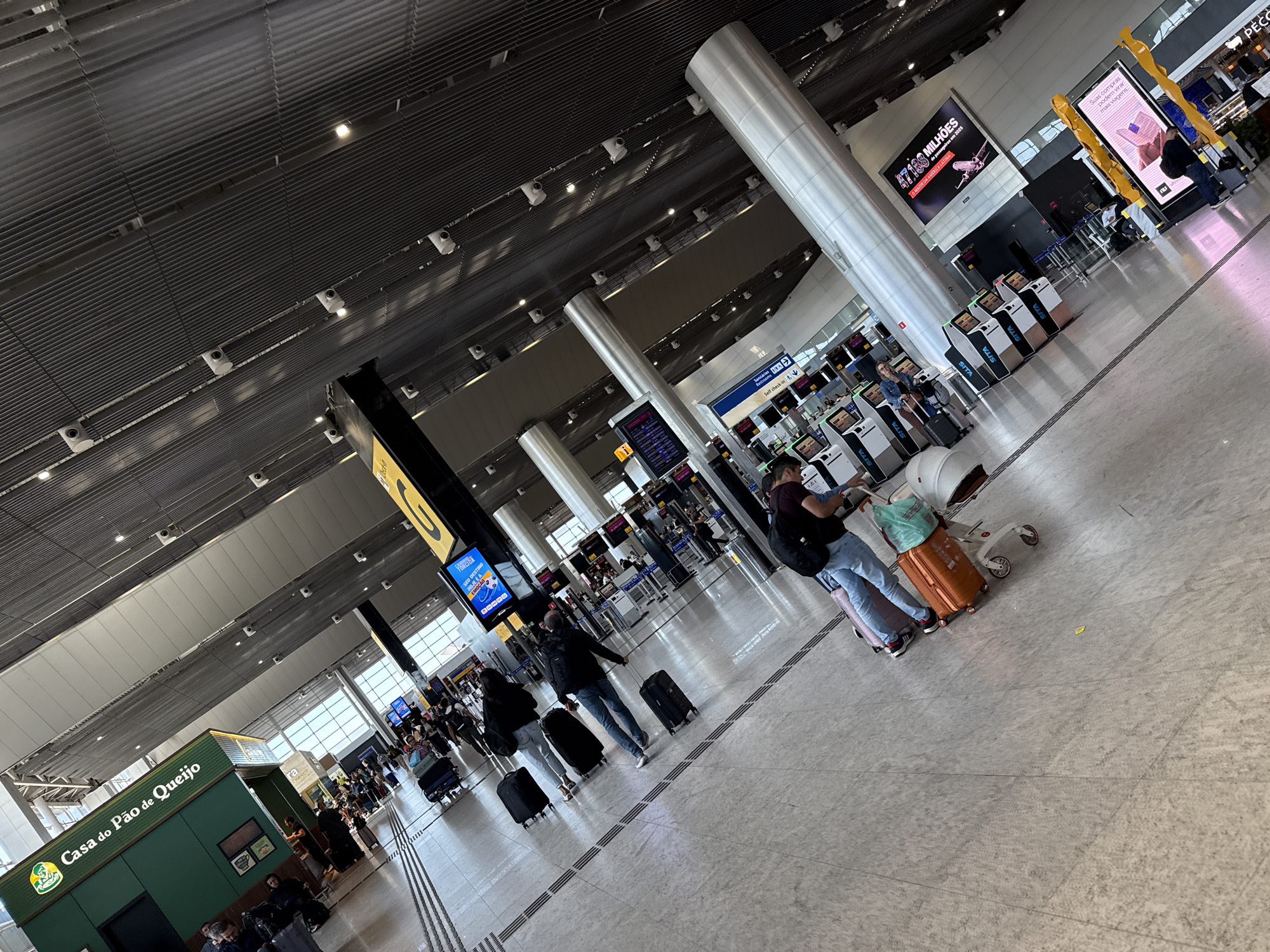 The image depicts an indoor scene within a departure hall at Guarulhos International Airport (GRU) in Guarulhos, Brazil. The perspective is a high-angle diagonal view, looking across the hall towards a series of check-in counters and a retail establishment.
The environment features a high, dark, corrugated metal ceiling equipped with numerous small recessed lights and white surveillance cameras. Several large, cylindrical metallic pillars support the ceiling structure. Natural light enters the space through large windows visible in the background.
In the upper-left quadrant, a prominent green kiosk with white lettering identifies it as "Casa do Pão de Queijo" (House of Cheese Bread). Below this, multiple individuals, presumably travelers, are present. In the mid-ground, a woman wearing light-colored pants pulls two black wheeled suitcases. Ahead of her, a man in a dark jacket and blue jeans pulls a single black wheeled suitcase. In the foreground, a man wearing a dark top and light blue jeans pulls a pink wheeled suitcase and a large orange wheeled suitcase, while simultaneously pushing a white baby stroller that carries additional items.
Along the right side of the frame, a long row of self-check-in kiosks, many branded "SITA," are visible, with some individuals interacting with the screens. Large digital display screens are mounted on pillars; one displays "Seu destino" (Your destination) and another advertises "47.168 MILHÕES de passageiros em 2025" (47.168 MILLION passengers in 2025). An overhead black sign with a large yellow "C" is positioned above the kiosks. The floor is a light-toned, speckled tile or concrete surface marked with parallel dark lines, possibly indicating pathways or queue boundaries.