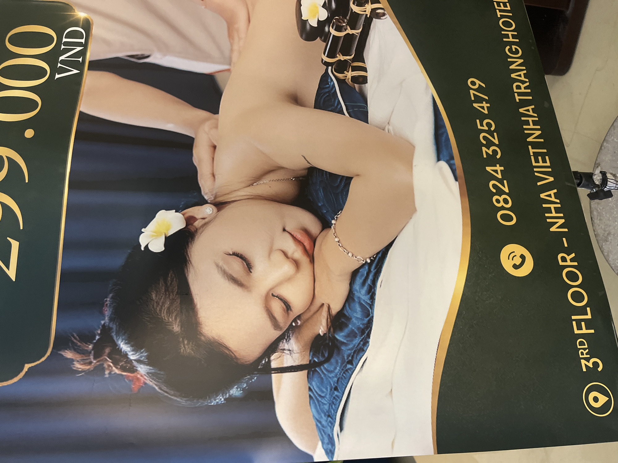 The image is a close-up of a poster advertising a spa. A woman with her eyes closed and a flower in her hair is lying face down on a massage table, receiving a massage from another person whose hands are visible. To the right of the woman are stacked massage stones and bamboo sticks. The bottom of the poster displays a phone number, "0824 325 479", and an address, "3RD FLOOR - NHA VIET NHA TRANG HOTEL". The poster has a dark green background with gold trim. The setting is in an unknown location in the city of Unknown, Unknown.