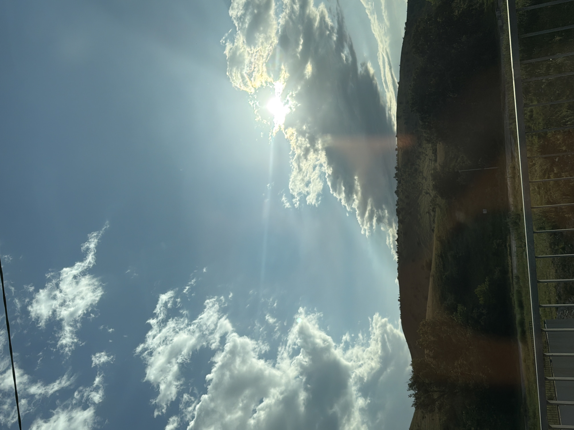 The image depicts a sunlit sky with scattered clouds. The sun is partially obscured by a large cloud formation, casting rays of light through the atmosphere. In the background, a treed hillside rises, with a paved path or road running along its base. To the right of the hillside, a metal railing with vertical supports is visible, suggesting the image might have been taken from an elevated position or next to a structure. The overall scene suggests an outdoor setting during the daytime, likely a clear to partly cloudy day. The orientation of the image is rotated 90 degrees counter-clockwise. There is no visible text in the image. The location context provided is Новая Джегута, Russia.