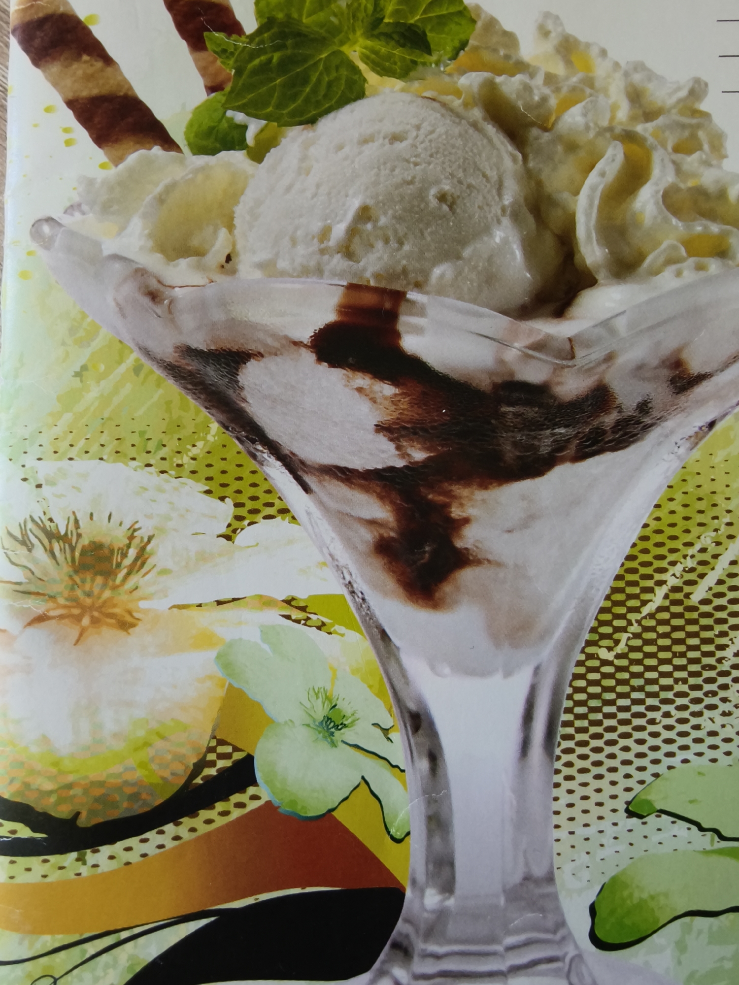 The image depicts an ice cream sundae in a clear glass goblet. The sundae consists of vanilla ice cream, whipped cream, chocolate syrup, a wafer stick, and a sprig of mint. The goblet rests on a surface with a patterned background featuring stylized flowers and abstract designs. The setting is not explicitly identifiable as Kryvyi Rih, Ukraine, as there are no specific landmarks or indicators.