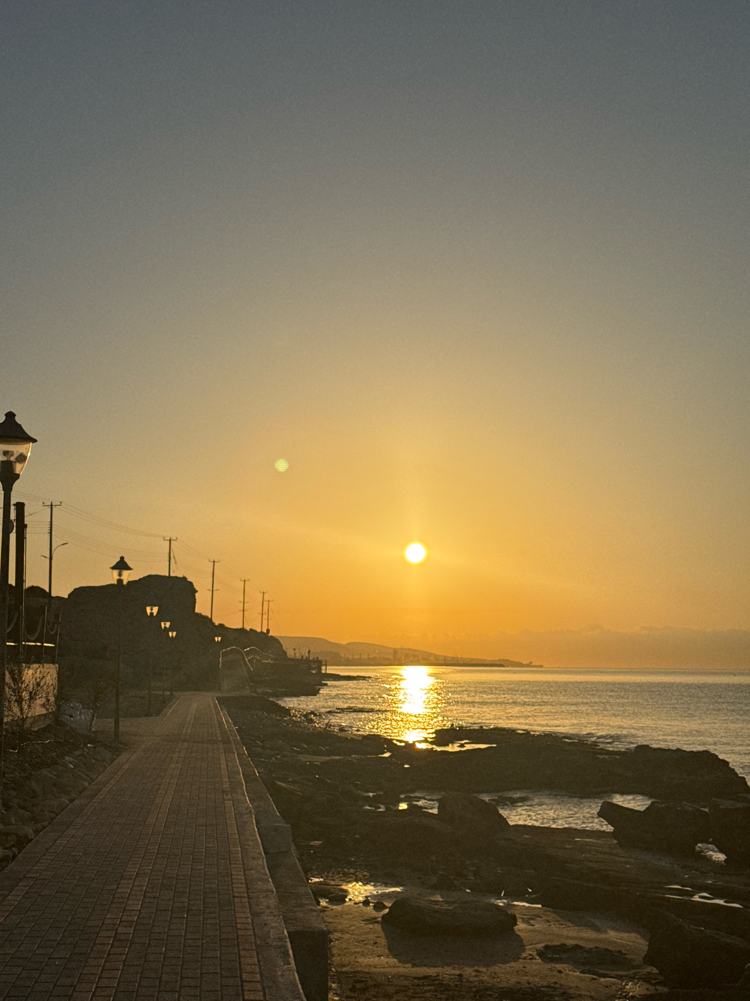 The image depicts a sunset scene along a coastal walkway in Agios Tychona Community, Cyprus. The sun is low on the horizon, casting a warm golden glow across the sky and sea. The walkway, paved with reddish-brown bricks, runs along the left side of the frame, bordered by a low stone wall and rocky terrain. Several lampposts are visible along the walkway and on the elevated rocky area to the left. The sea is calm, with gentle waves lapping against the dark, rocky shoreline. The distant coastline is visible, with the silhouette of hills and possibly buildings. The overall atmosphere is serene and peaceful.