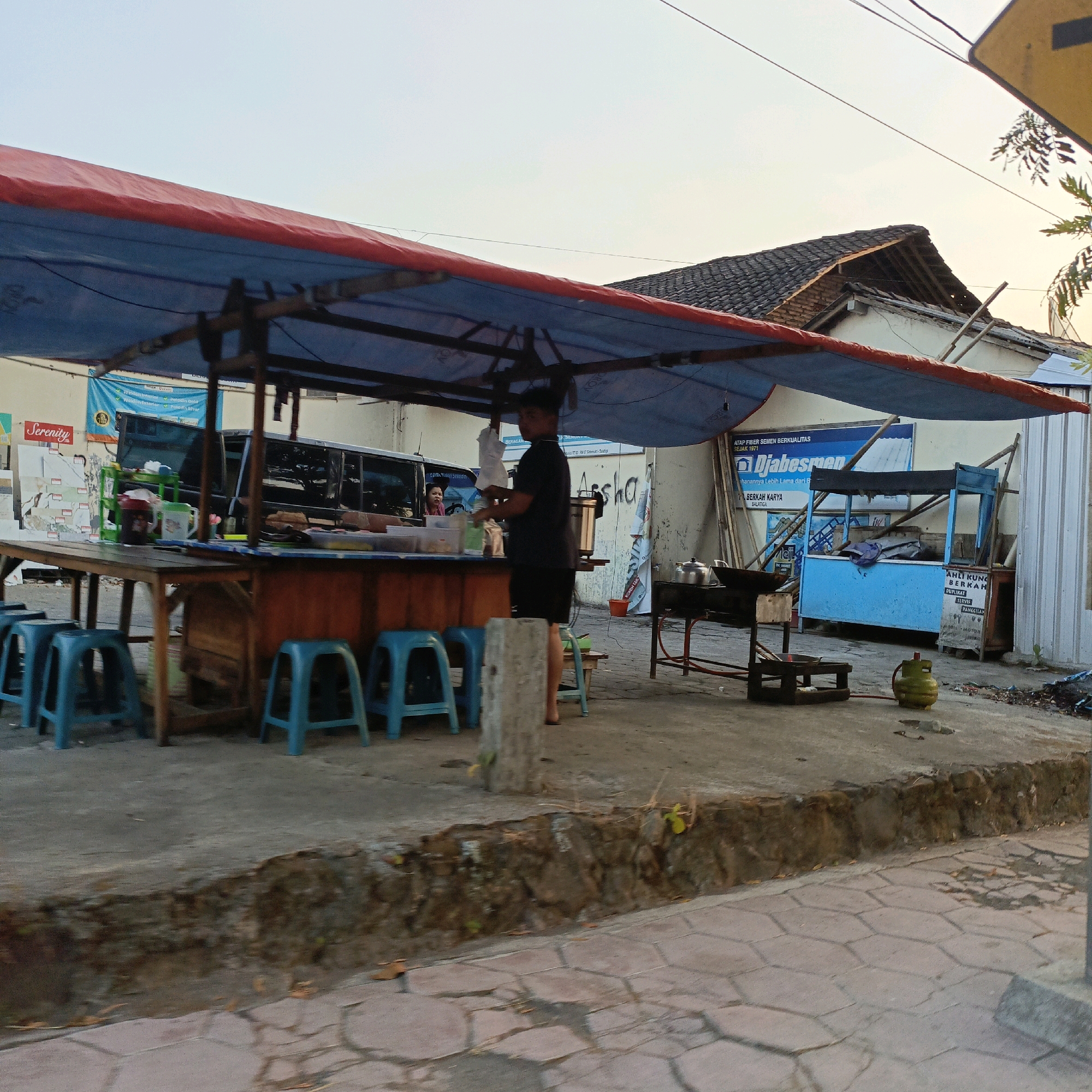 This image depicts a street scene in Salatiga, Indonesia, likely in the late afternoon or early evening, indicated by the soft, warm lighting. The central focus is a food stall set up under a blue and red tarp supported by wooden poles. A man wearing a dark t-shirt and shorts is standing behind the stall, engaged in an activity related to food preparation, possibly serving a customer or arranging items on the counter. To the left, several teal plastic stools are lined up in front of the stall, suggesting a seating area for patrons.
Behind the stall, a dark-colored vehicle is visible, partially obscured by the awning. The stall itself is constructed with a wooden counter and what appears to be a wooden cabinet or storage unit below it. Various food containers and utensils are present on the counter. Further to the right, another small food cart, painted blue, is positioned. It is empty, and a green gas cylinder sits in front of it. A small cooking stove with a pot on it is also visible to the right of the main stall.
The background includes a building with a tiled roof and a light-colored wall. Several signs are affixed to the wall, including one with text that appears to be in Indonesian, advertising products or services such as "Djabesmen" and "ATAP FIBER SEMEN BERKUALITAS". The ground is paved with concrete and some areas show signs of wear and tear, with a low stone wall bordering a section of the foreground. The overall environment suggests a typical roadside food vendor setup in an urban or semi-urban setting.
