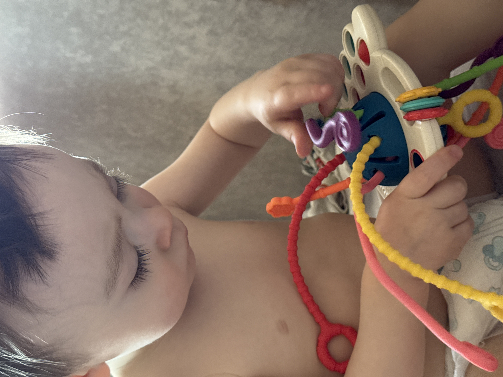 A baby is lying on their back, holding a colorful toy. The toy is made of white plastic with several holes and colorful rubbery strings protruding from the holes. The baby is grasping one of the strings with their left hand and holding the toy with their right hand. The baby is fair-skinned with dark hair. The baby is wearing a diaper. The background is a textured surface. There is no indication of the location being Izhevsk, Russia.