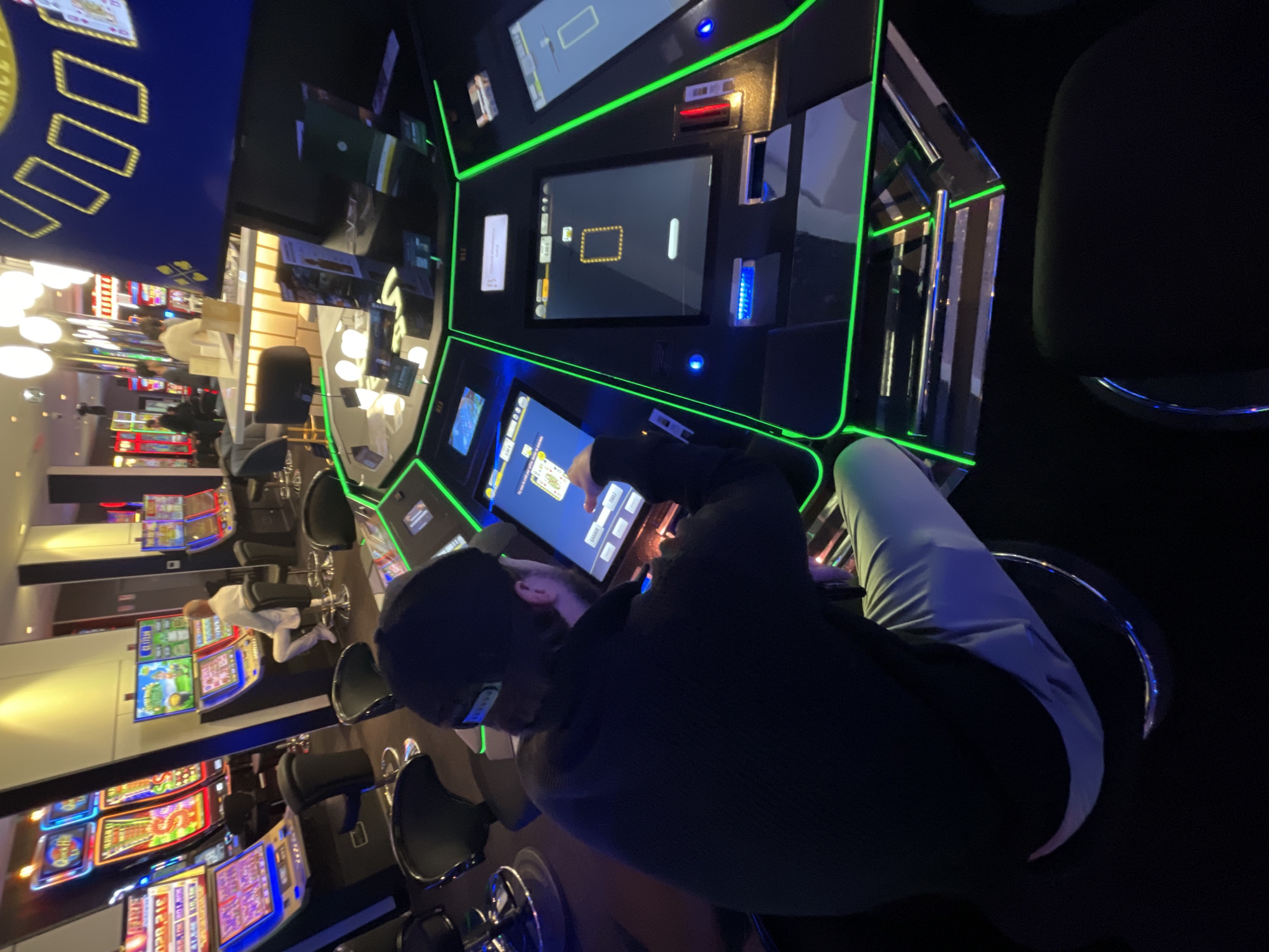 An individual is depicted from behind, seated at a multi-station electronic gaming table within what appears to be a casino, set in the evening or night due to the dim ambient lighting and prominent artificial illumination. The main subject wears a dark long-sleeved shirt and light-colored pants, their hands engaged with a touchscreen display, playing a card game with visible controls like "BET," "DEAL," "STAND," and "HIT."
The gaming table is outlined with striking neon green lights and features several screens. Adjacent screens display a waiting interface with a golden rectangular outline. In the background, a vibrant array of brightly lit slot machines line the walls, and another person in white is visible interacting with them. The atmosphere is one of focused entertainment, dominated by the glow of electronic displays. Numerical identifiers, such as "218," are present on the gaming station panels. Faint text hinting at "FRANCE" is visible on an overhead sign. This scene captures a moment of play in a modern gaming establishment, likely situated in Sainte-Maxime, France.