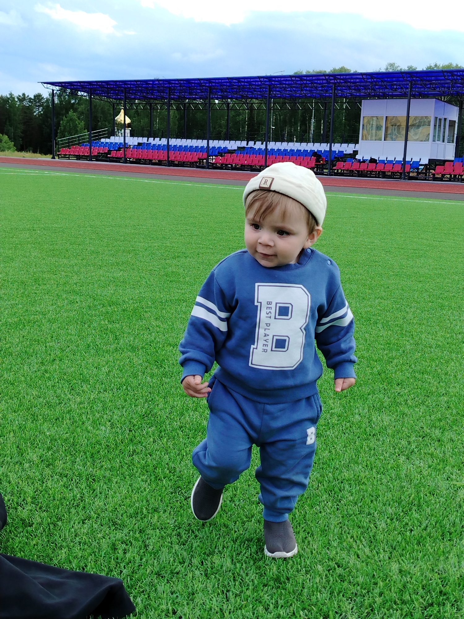 A toddler stands on a green field wearing a blue tracksuit, gray shoes, and a cream-colored cap with a letter "R" on it. The toddler is moving to the left. In the background, there is a stadium with red, white, and blue seating, and a small white building. Trees are visible behind the stadium. The sky is overcast. The location is Noviy, Russia.
