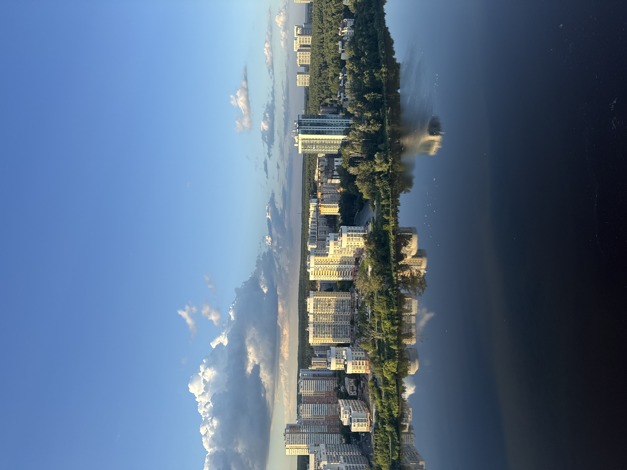 This is a high-angle, full-shot photograph of a cityscape overlooking a body of water. The sky is a clear blue with a few scattered white clouds, illuminated by what appears to be late afternoon sunlight, casting warm tones on the buildings.
In the middle ground, a densely populated residential area lines the edge of the water. Numerous multi-story apartment buildings, with varying architectural styles and colors, dominate the scene. Some buildings are tall and slender, while others are wider and more block-like, featuring balconies and a mix of light and darker colored facades. A dense forest of green trees serves as a backdrop to these buildings, extending further into the distance.
The foreground is occupied by a large, dark body of water, likely a river or lake, which reflects the buildings and the sky above. The reflections are somewhat distorted but clearly visible, adding depth and symmetry to the image. The surface of the water is mostly calm, with some subtle ripples.
The location is identified as the Yekaterinburg-City area, specifically near the Iset River, in Yekaterinburg, Russia. The image captures the urban development and natural landscape of the city. There are no people or animals visible in the frame. The overall impression is one of a modern, developing city integrated with its natural environment.