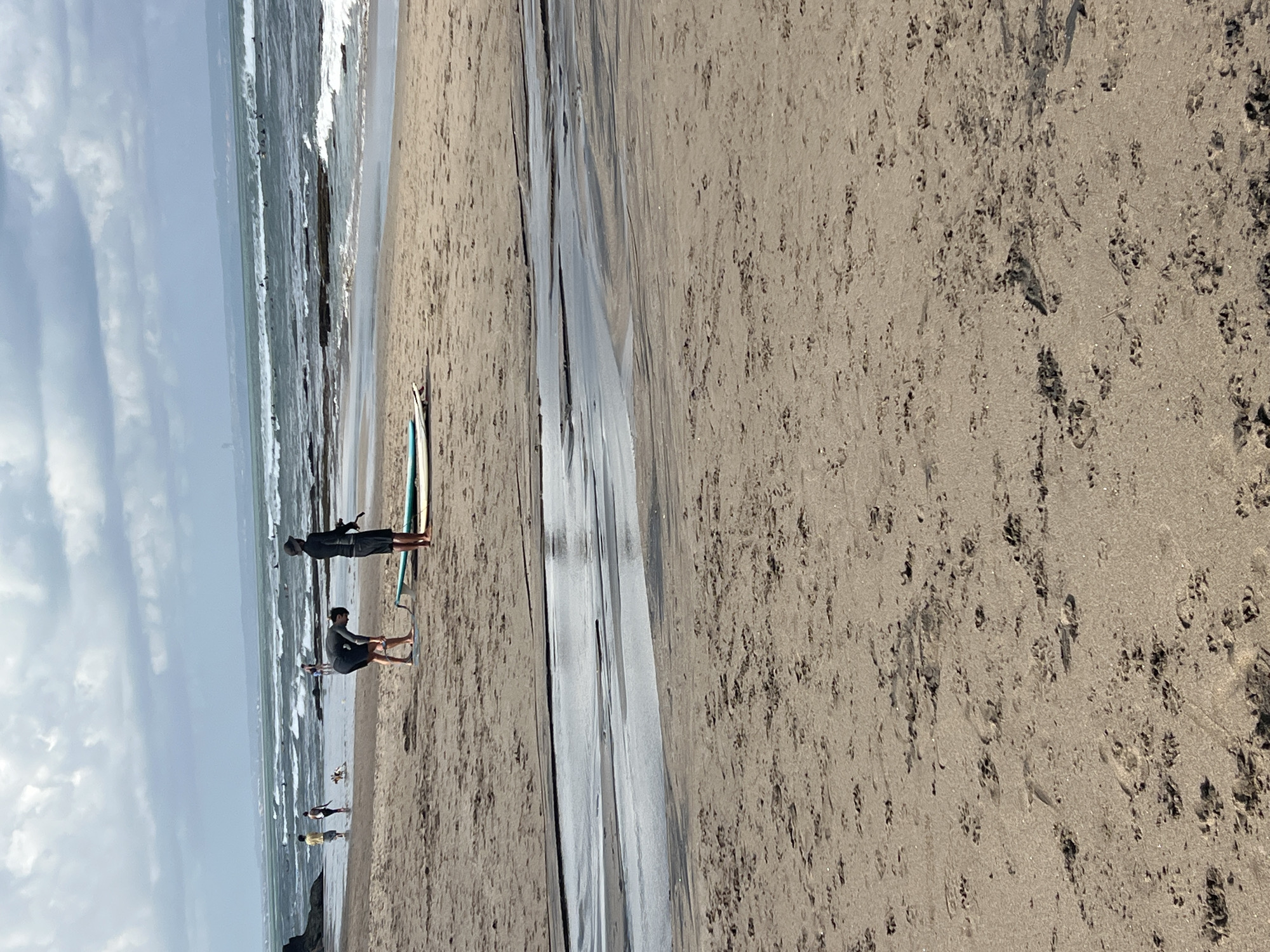 A moderately cloudy day at a beach in North Kuta, Indonesia. The foreground shows a sandy beach with footprints and a shallow stream of water. In the mid-ground, two men are standing on the sand. One man, wearing a dark t-shirt and shorts, is bending over near a surfboard. The other man, dressed in black and wearing a hat, is holding a surfboard and looking towards the water. Further back, several other people are visible on the shore and in the ocean. The ocean has moderate waves, and there are surfers in the water. The background shows the coastline and a hazy horizon.
