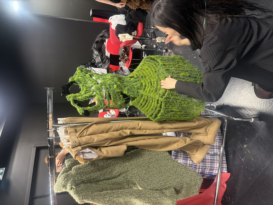 The scene depicts a backstage or studio area in Paris, France, characterized by black walls and a dark floor. A chrome rolling clothing rack is positioned centrally, holding multiple garments. On the left, a voluminous olive-green, shag-textured coat or sweater hangs. To its right, a pair of light brown tailored trousers is displayed. Dominating the right side of the rack is a sculpted, sleeveless garment resembling a top or dress, entirely covered in intricate bright green, moss-like material with a textured, drooping appearance.
In the foreground, a woman with long dark hair, wearing a black pinstriped blazer, kneels or squats, meticulously adjusting the lower portion of the moss-like garment with her right hand. Her left arm is slightly raised. In the upper left background, another individual with dark hair is partially visible, appearing to attend to items near the top of the clothing rack. The background features several red chairs, some laden with discarded clothing or plastic bags, and a black trash bag on a surface, indicating a functional, possibly chaotic, workspace. A blue and white plaid bag rests on the floor beneath the clothing rack.