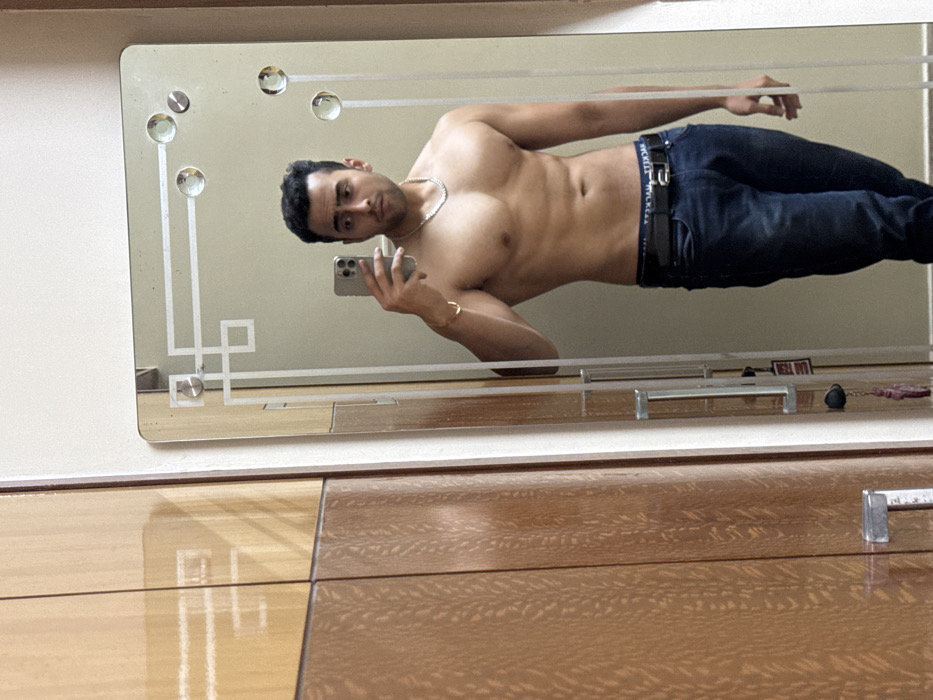 An image displays a full-length mirror selfie of a man, rotated 90 degrees counter-clockwise. The man, appearing horizontally oriented within the frame, is shirtless, showcasing a muscular torso. He wears dark blue jeans, a black belt with a partially visible "WICKERLY" brand name on the buckle, a silver chain around his neck, and a gold bracelet on his left wrist. He is holding a smartphone to take the picture, which is reflected in the mirror.
The mirror itself is rectangular and features decorative etched patterns, including geometric lines and shapes, and several circular glass embellishments along its top and left edges. The background reflected in the mirror appears to be a plain, light-colored wall.
Below the mirror, and forming the foreground of the image, is a wooden surface, likely a dresser or table, with a glossy finish and a distinct wood grain pattern. Reflected on this surface, within the lower part of the mirror, are a couple of small objects, including what looks like a metallic handle or pull, and a small, dark spherical object possibly attached to a chain. A small white label with unclear red text, possibly reading "DELL OUT", is also visible in this reflection. The man maintains a neutral to slightly serious expression. The scene is indoors.