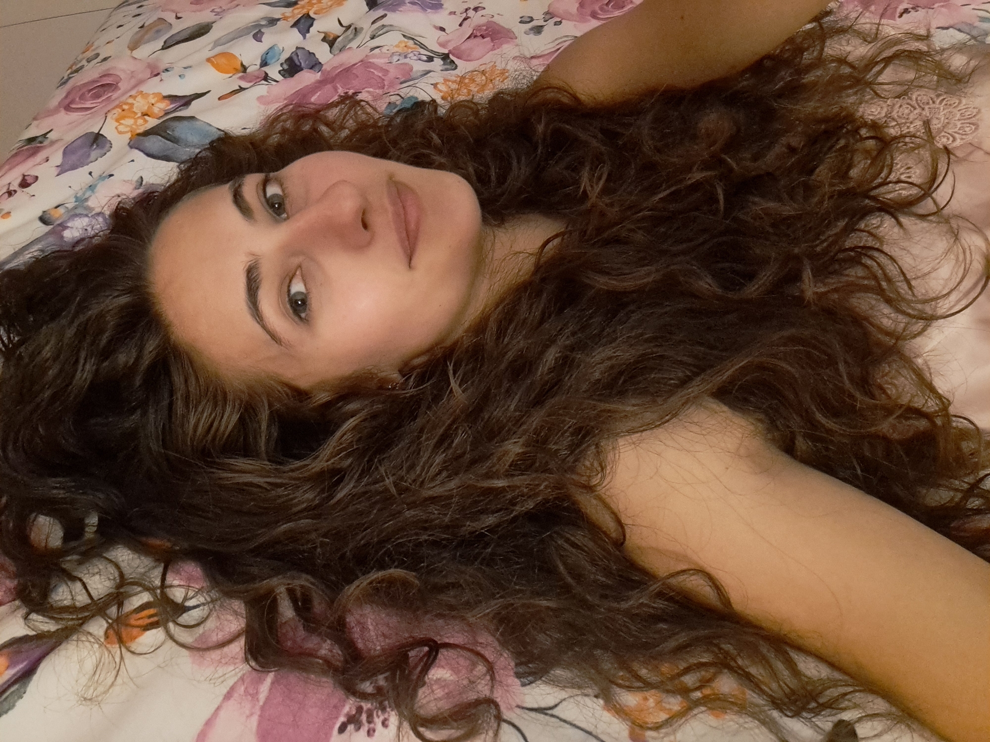 A young woman with long, dark, curly hair is lying on a bed with floral-patterned sheets. She is looking up at the camera with a slight smile. Her hair is spread out around her head and shoulders. The sheets are white with pink, purple, and orange floral designs. The woman is wearing a light pink garment with lace trim. The scene appears to be indoors, possibly in a bedroom. There are no other people or objects of significance visible in the image. The lighting is soft and natural, suggesting daylight.