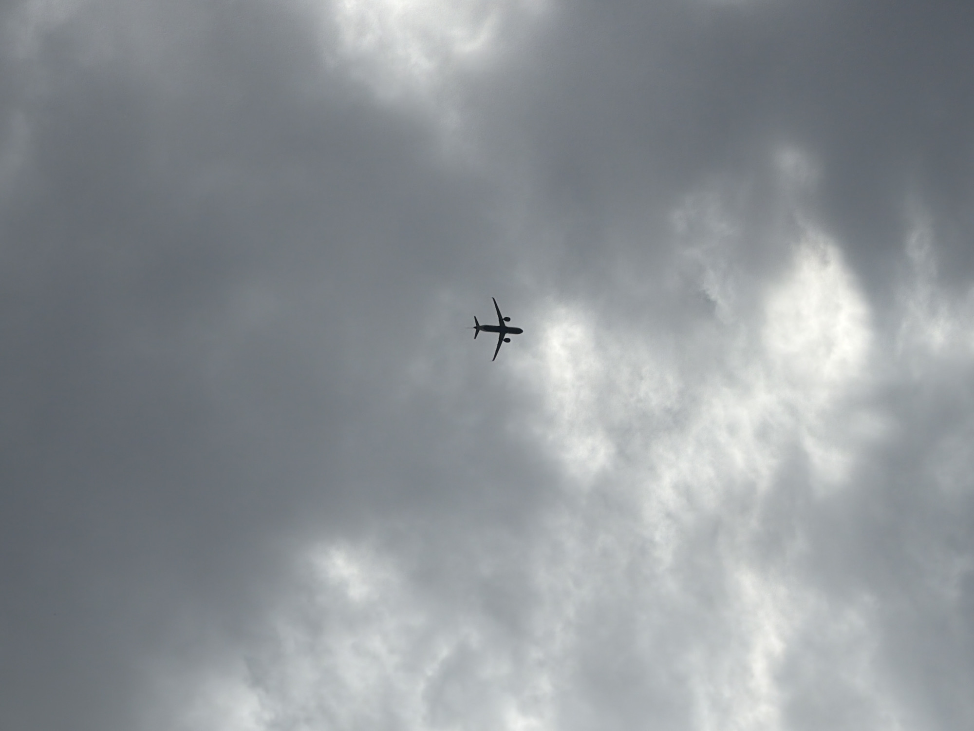 A dark silhouette of an airplane flies overhead against a cloudy sky. The location and date are unknown, but it's possibly Sant Martí Sarroca, Spain.
