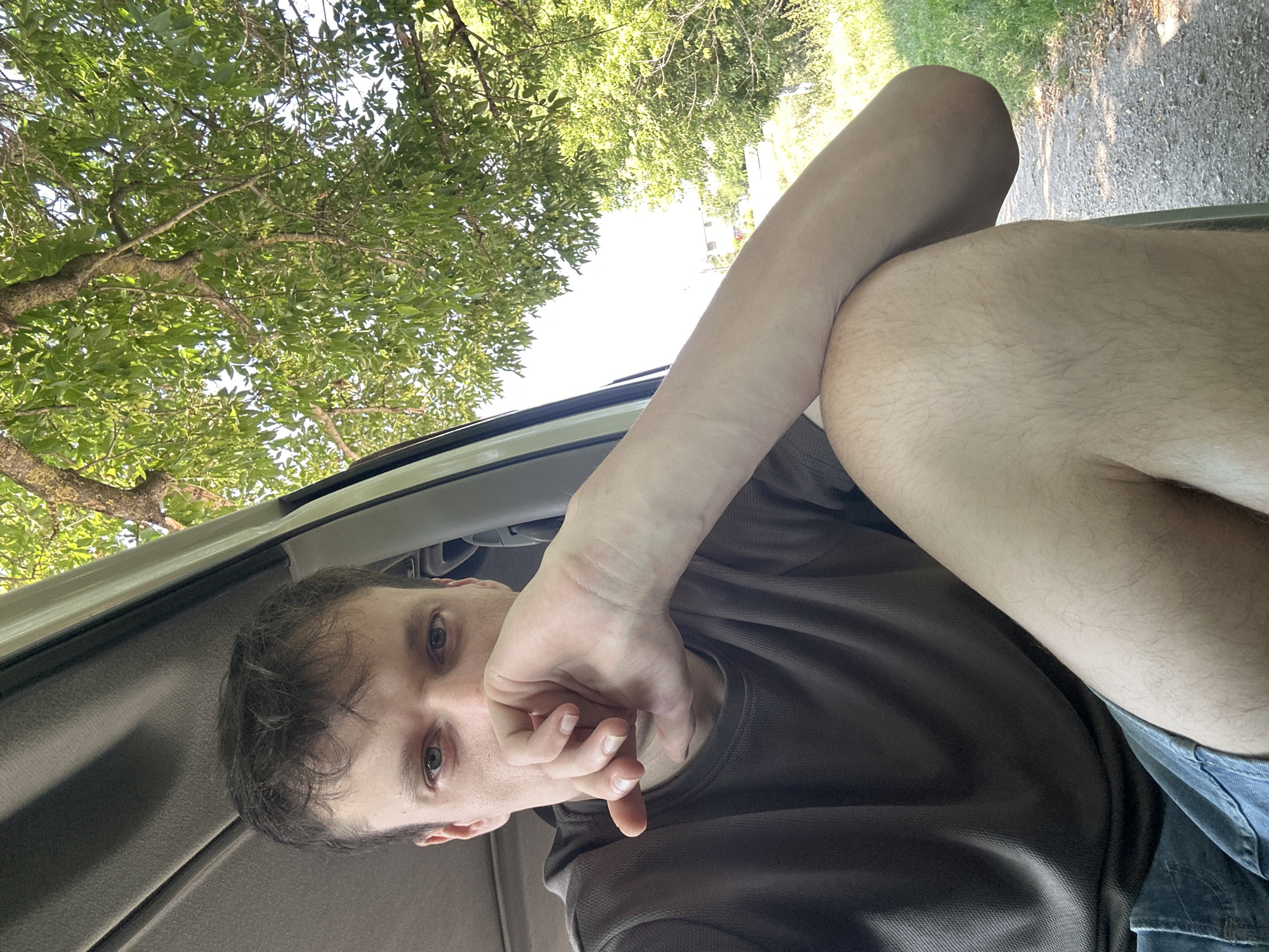 A young man with short brown hair and blue eyes is captured in a medium close-up, seated inside a vehicle with the door open. His right hand is brought to his face, resting on his chin, with his fingers covering his mouth. He is wearing a dark grey t-shirt and denim shorts. The interior of the vehicle, likely a van or SUV, is visible, with a grey headliner and frame of the open door. Outside, lush green trees with dappled sunlight filter through, suggesting a rural or semi-rural setting. The background shows a gravel path and more greenery, indicative of a natural environment. Based on the provided context, the location is Makiv, Ukraine.