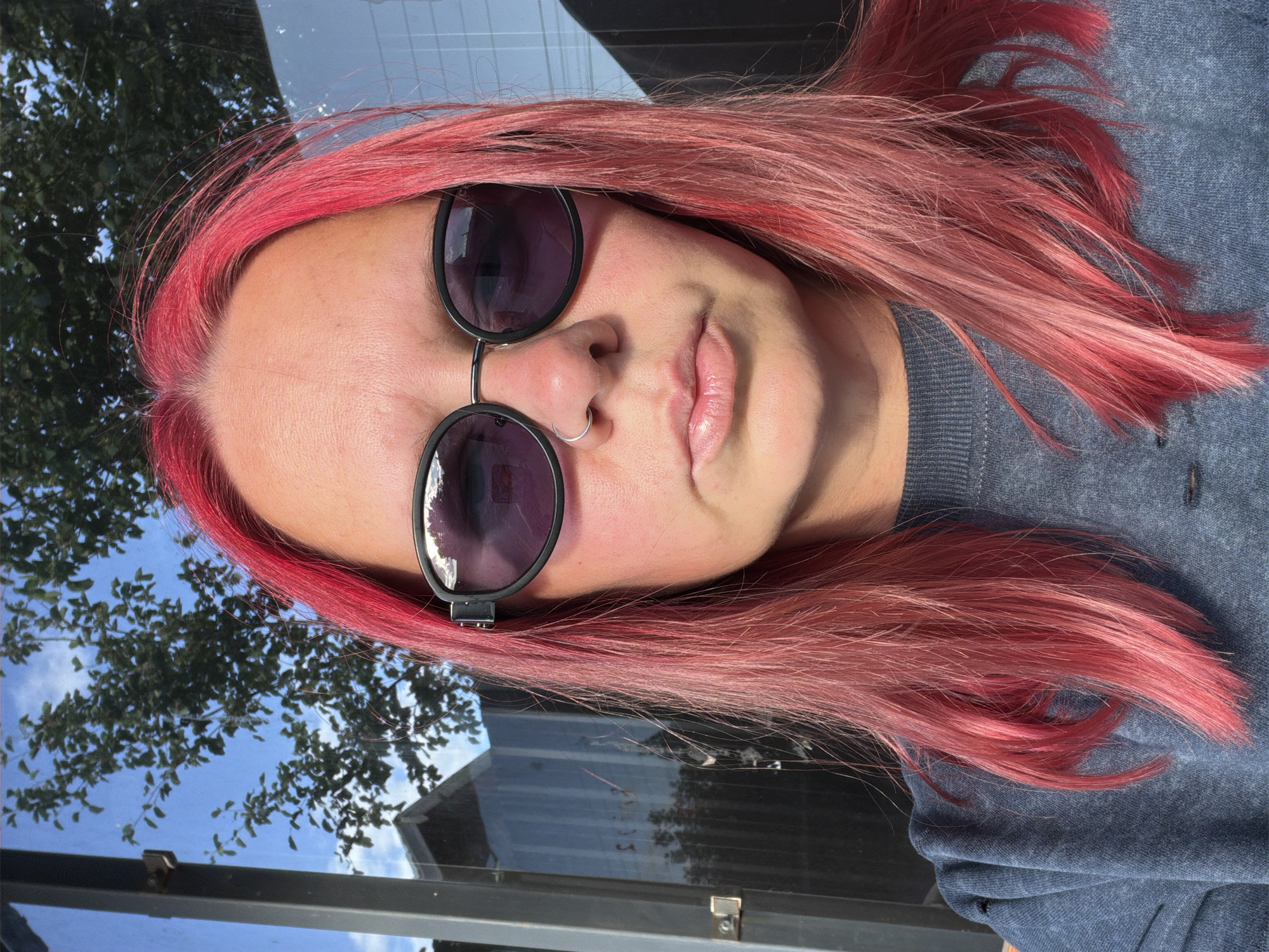 A woman with bright pink hair and sunglasses poses for a selfie outdoors. She is wearing a dark blue sweatshirt. Behind her, a tree with green leaves is visible against a blue sky with some clouds. The background also shows a building with glass windows and a metal frame. The scene appears to be on a sunny day.