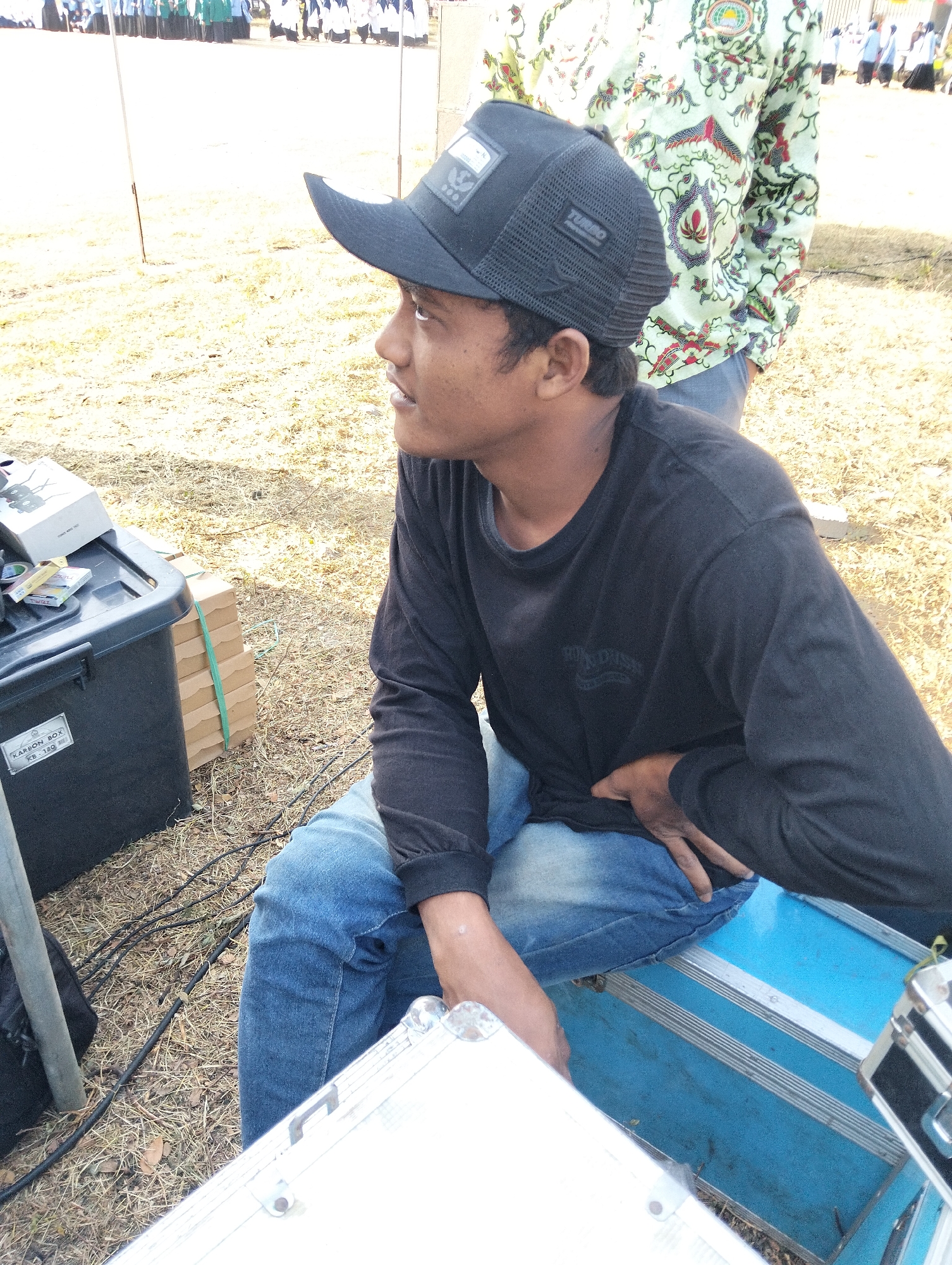 An outdoor daytime medium shot shows a young man wearing a black long-sleeved shirt, blue jeans, and a black baseball cap, sitting on a blue metal case. He is looking to his left, with his right hand resting on his lap. The background shows dry grass, some black cables, a black plastic box labeled "KARBON BOX KB 180," and a stack of cardboard boxes. In the far background, a group of people in uniforms stand in formation. The scene appears to be an outdoor event in Paciran, Indonesia.