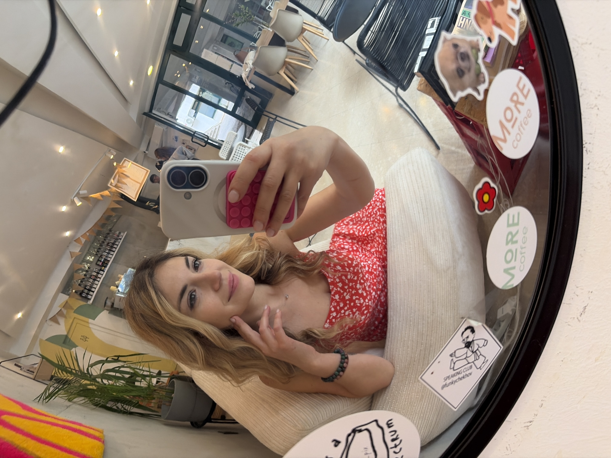 A woman is taking a selfie in a mirror. The mirror is in a cafe or restaurant setting in Budva, Montenegro. The woman has blonde hair and is wearing a red dress. She is holding a phone with a pop socket on the back. The mirror has stickers on it, including one that says "MORE coffee". The reflection shows other patrons seated at tables and chairs. There are plants in the foreground. The decor includes pennants and decorative bottles on shelves. The setting is indoors.