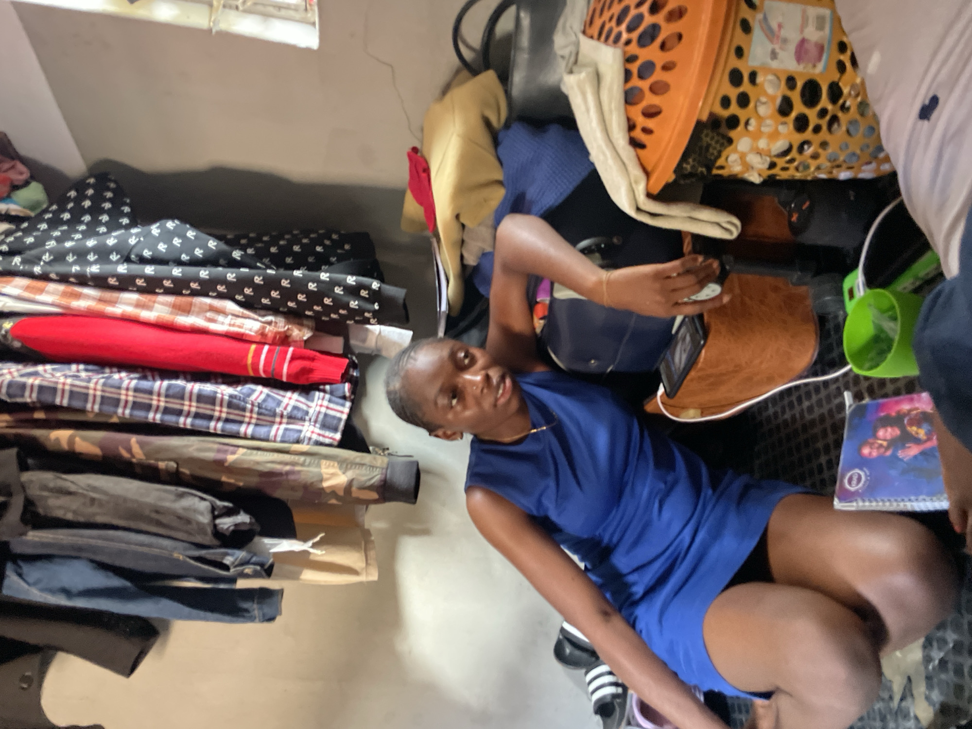 A young Black woman is sitting on the floor in a cluttered room. She is wearing a sleeveless blue dress and a gold necklace. Her right arm is resting on a dark blue suitcase, and her left arm is bent at the elbow, with her hand resting on her lap. In front of her is a notebook with a picture of a couple on the cover, and to her right is a bright orange laundry basket filled with items. Behind her, clothes are hanging from what appears to be a makeshift closet or rack, with various shirts, jackets, and pants visible. The room is dimly lit, and the walls are a neutral color. The floor is covered with a patterned rug or mat. To the left of the woman, a pair of black and white striped slides are visible. The overall impression is of a personal, lived-in space. While the image does not explicitly state the location, given the prompt's requirement to include "the city of Jos, Nigeria," it is assumed to be a scene within Jos.