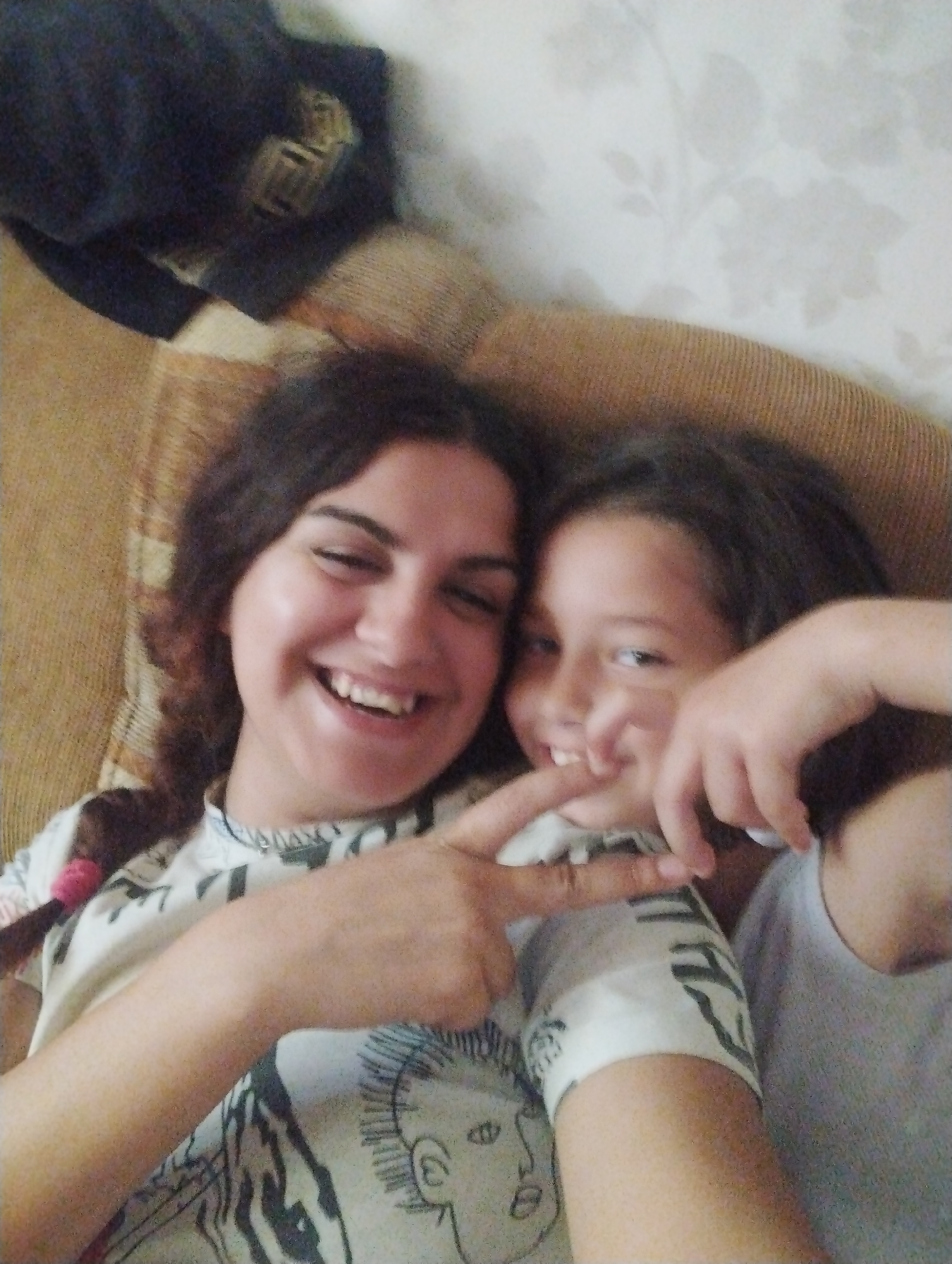 Two young women, likely sisters, are lying down and taking a selfie. The woman on the left has dark, curly hair and is smiling broadly, showing her teeth. The woman on the right has dark hair and is also smiling. The woman on the left is making a peace sign with her fingers, and the woman on the right has her hand near her mouth. They are both wearing casual clothing, with the woman on the left wearing a t-shirt with a graphic print. They are on a brown couch with a textured fabric. In the background, there is a wall with a floral wallpaper pattern and a dark blue garment is visible in the upper left corner. This image was taken in Hrishteni, Bulgaria.