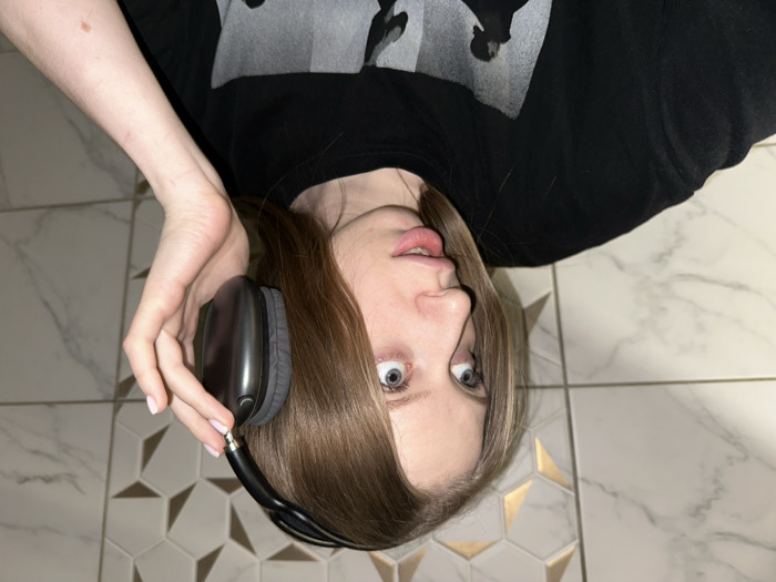 A young female, characterized by long, straight, light brown hair and blue-grey eyes, is captured in a close-up, chest-up view. She is looking upwards and to the right of the frame with a slightly open mouth, conveying an attentive or surprised expression. She wears a black short-sleeved t-shirt featuring a grey and white graphic print, partially visible on the torso. Her right hand, adorned with light pink nail polish on the fingernails, grips the right ear cup of a pair of dark grey over-ear headphones, which are positioned against the side of her head. The background consists of an indoor wall covered in light-colored, possibly marble-patterned square tiles, with a distinct section of decorative hexagonal tiles featuring gold metallic accents interspersed with plain white hexagonal tiles. The precise location within the city of Chaikovsky, Russia, cannot be determined from the visual information provided.