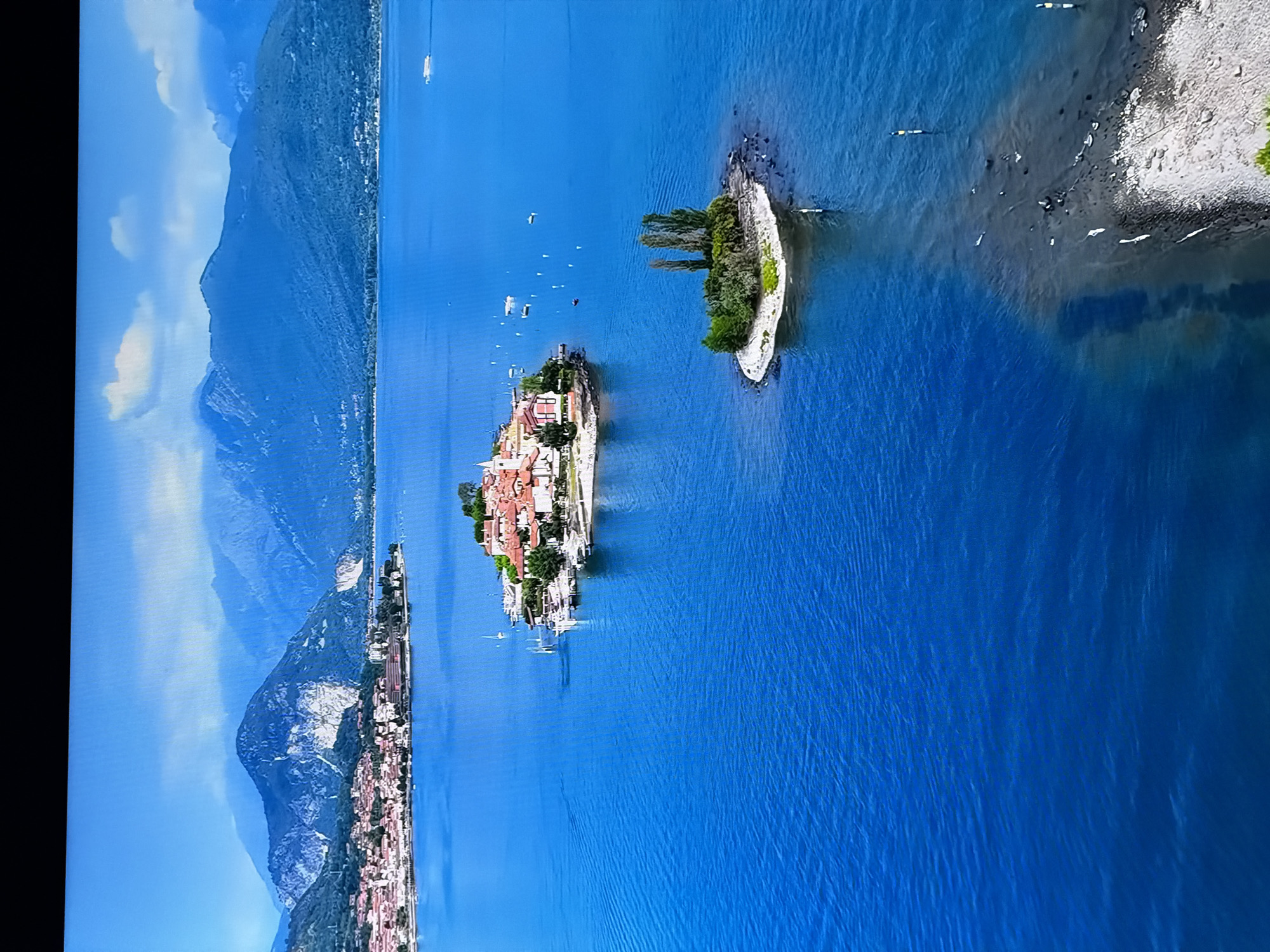 The image displays an aerial view of a serene lake with several islands. In the foreground, a shallow, rocky area extends from the bottom right into the water, with several white birds visible on the rocks and in the shallow water. Further into the lake, a small, circular island is covered with lush green vegetation and topped with three tall trees. Beyond this, a larger, more developed island features numerous buildings with red-tiled roofs, surrounded by greenery. Several boats are docked around this larger island, and more boats, including what appears to be a ferry, are scattered across the blue water. In the background, a picturesque town with terracotta roofs is nestled along the shoreline, backed by rolling green hills and majestic, mist-shrouded mountains under a clear blue sky with a few scattered white clouds. There are no indications of the city of Chirchik, Uzbekistan in this image.