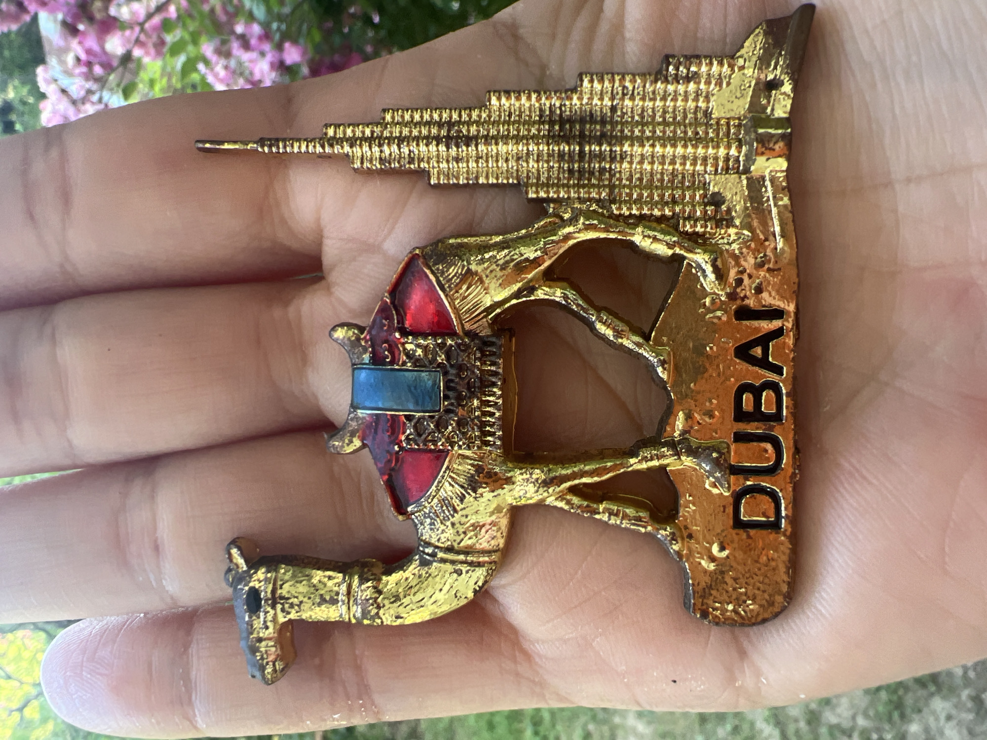 A close-up shot shows a golden metal souvenir magnet in the shape of a camel standing next to the Burj Khalifa building in Dubai. The camel is adorned with a red and blue saddle and is mounted on a base that reads "DUBAI" in black lettering. The souvenir is held in a person's hand, with their skin visible in the background. Out-of-focus pink flowers and green foliage can be seen in the background, suggesting an outdoor setting.