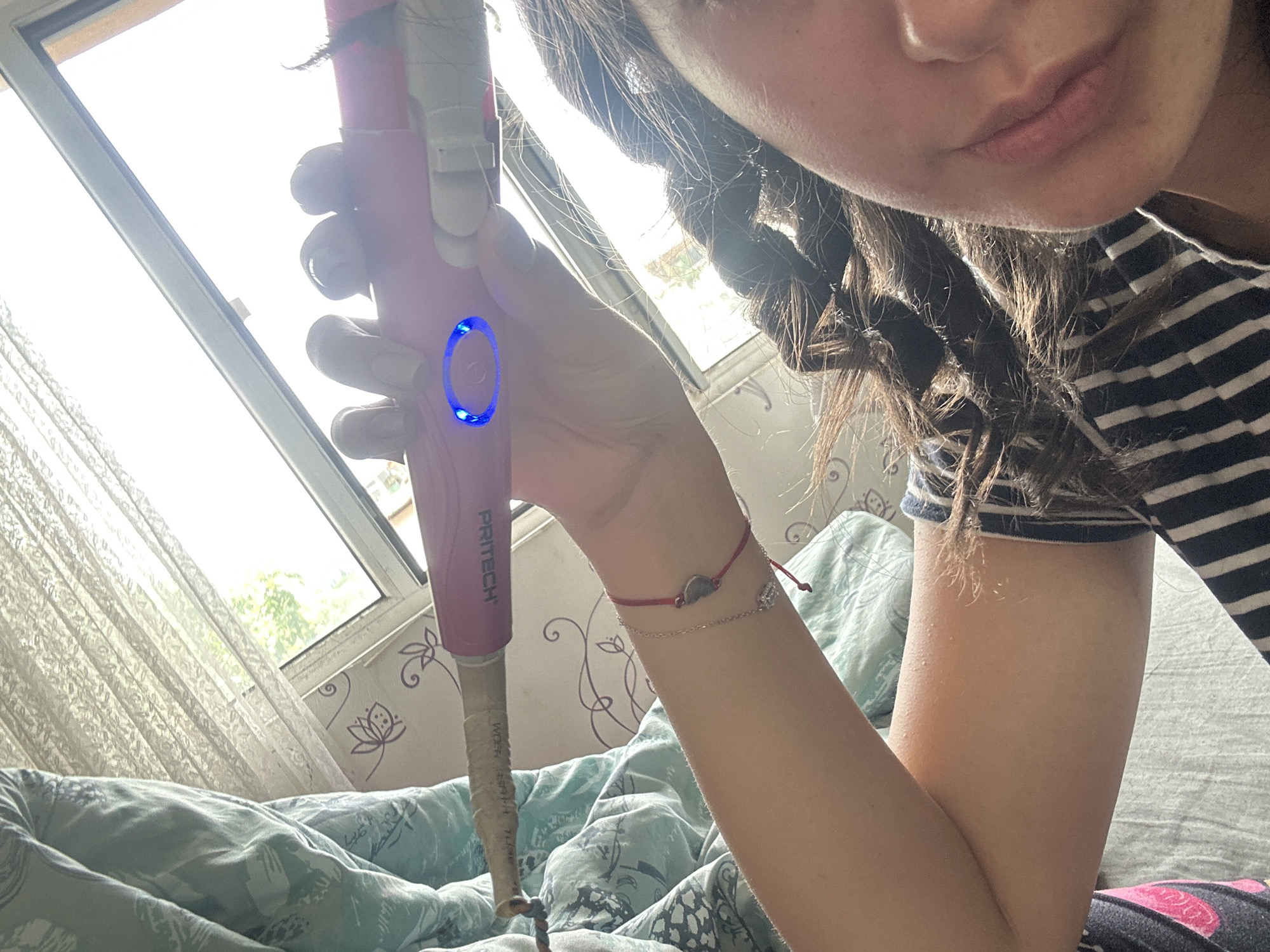A woman is holding a pink "PRITECH" branded hair curling iron in her left hand. The iron has a blue light illuminated. The woman has brown hair styled in braids. She is wearing a striped shirt. Her arm has two bracelets. She is in a room with patterned wallpaper and a window with sheer curtains. The bed has a blue and gray patterned duvet. The location in the city of Unknown, Unknown, is not known.