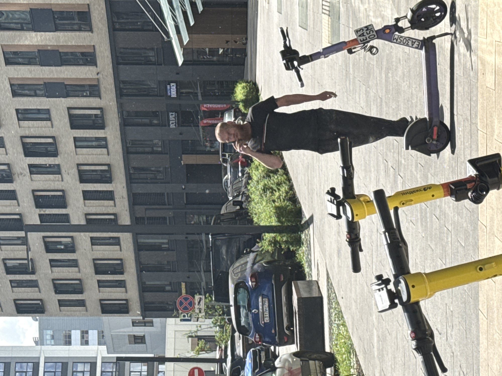 An urban street scene is captured in bright daylight, featuring a man, several electric scooters, parked cars, and modern buildings.
The central subject is a man with a shaved head and a beard, wearing a black t-shirt and dark pants. He stands on a paved sidewalk, holding a phone to his right ear and a lit cigarette in his left hand, appearing to be engaged in a phone conversation.
Around him, several electric scooters are parked. Two yellow scooters, identifiable by "ЯНДЕКС GO" (Yandex Go) branding visible on their handlebars, are in the foreground and midground. Further back, a purple electric scooter with a registration plate displaying "EA305M" is also parked.
In the background, a blue Renault Duster car with a Russian license plate "C317PC196" is parked near a patch of green bushes. Other vehicles are partially visible behind it. Two multi-story buildings dominate the background: one with light-colored brick or stone cladding and dark recessed windows, and an adjacent modern building with large, dark, reflective windows. Reflections of other buildings and sky are visible in these windows, including partial text like "БОРОДАЧ" (BORODACH) and "SAMSUNG".
A red circular 'no stopping' or 'no parking' sign is visible on a pole near the parked cars. The setting suggests a bustling city environment, consistent with Moscow, Russia, given the vehicle license plates and scooter branding. The time of day is clearly daytime, with ample natural light and soft shadows, indicating good weather.
